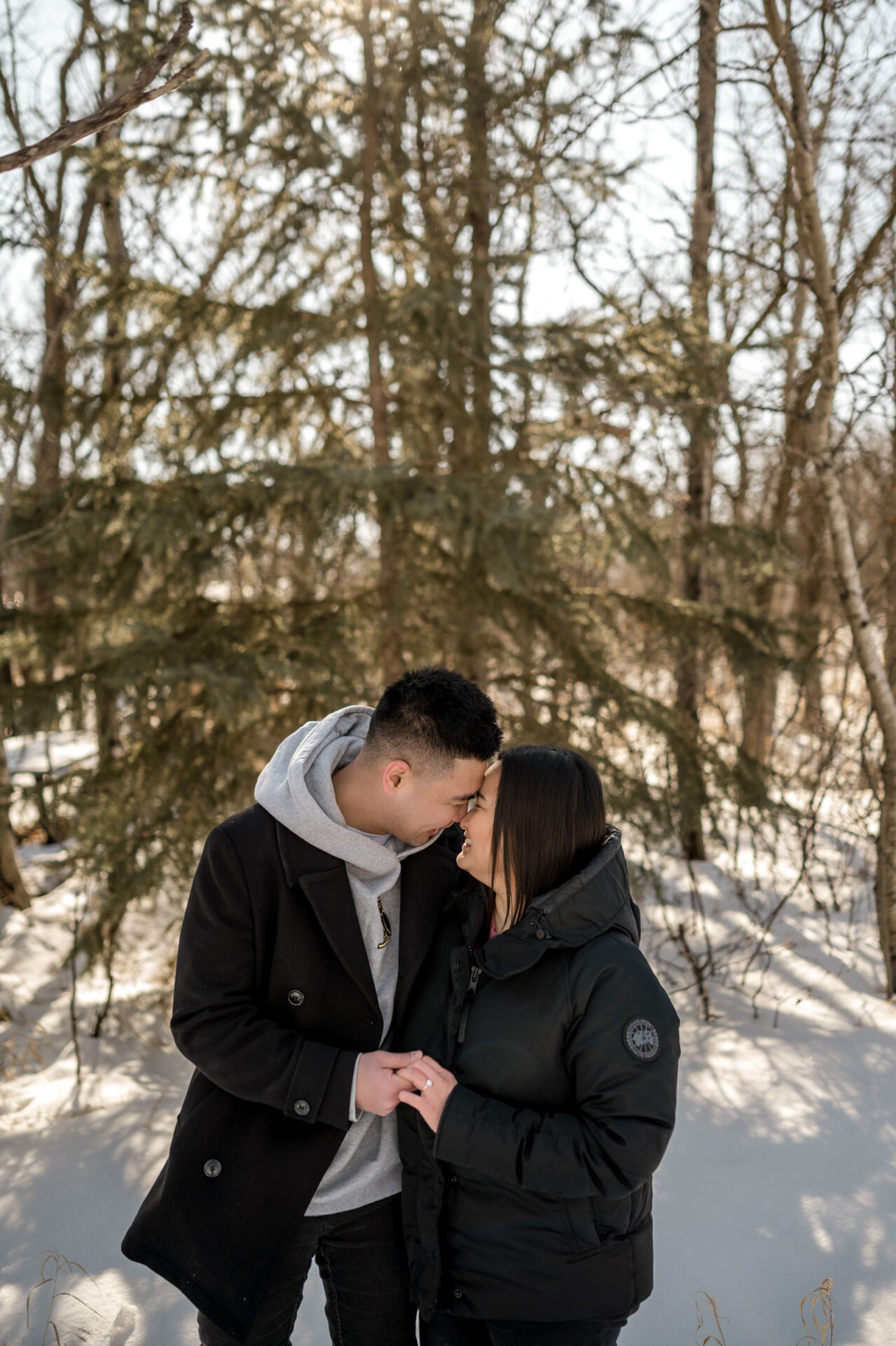 Winter engagement: couple in cozy attire, smiling closely in a snowy forest.