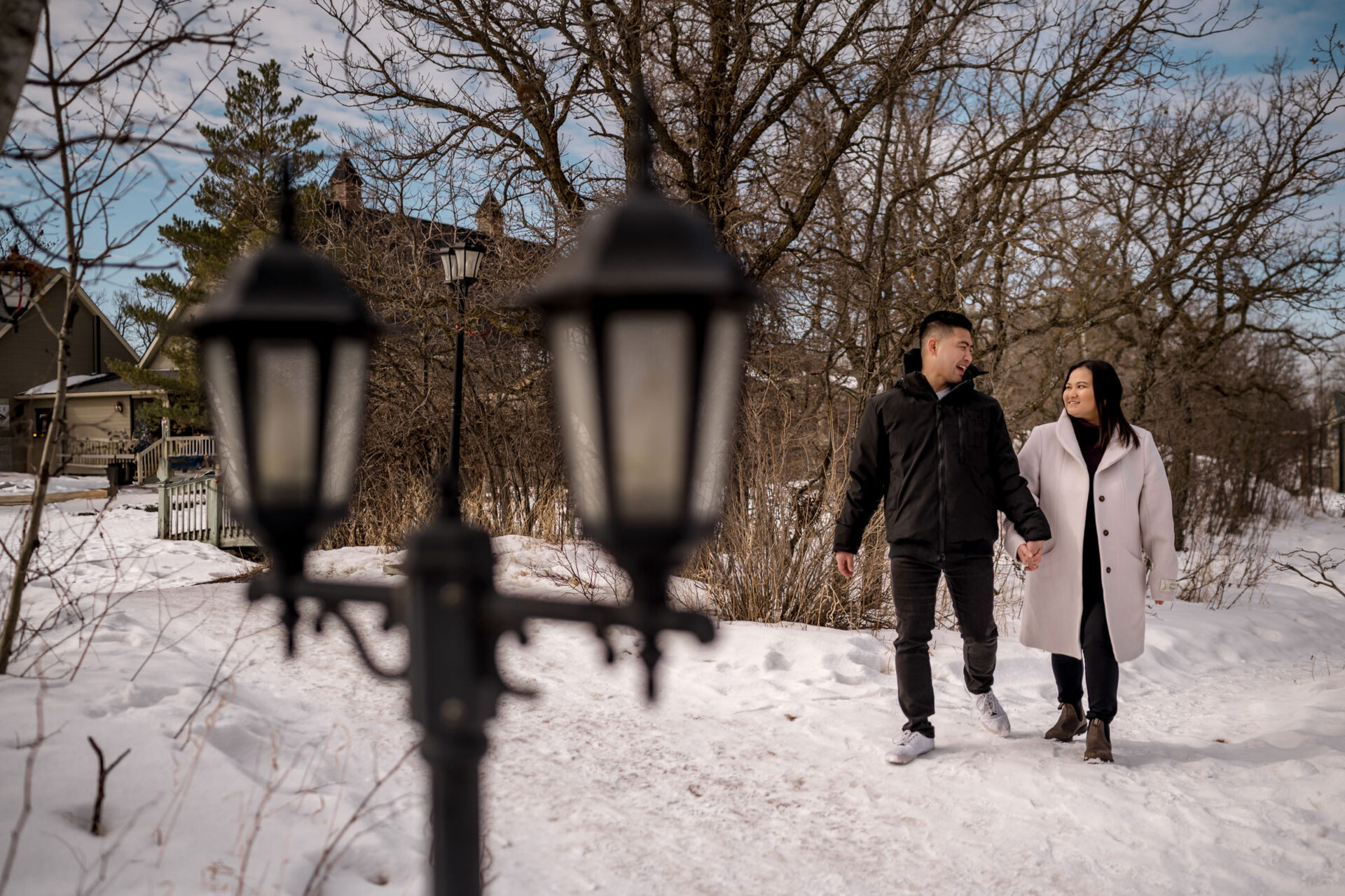 A couple holds hands, enjoying a winter engagement on a snowy path near a vintage lamp post.