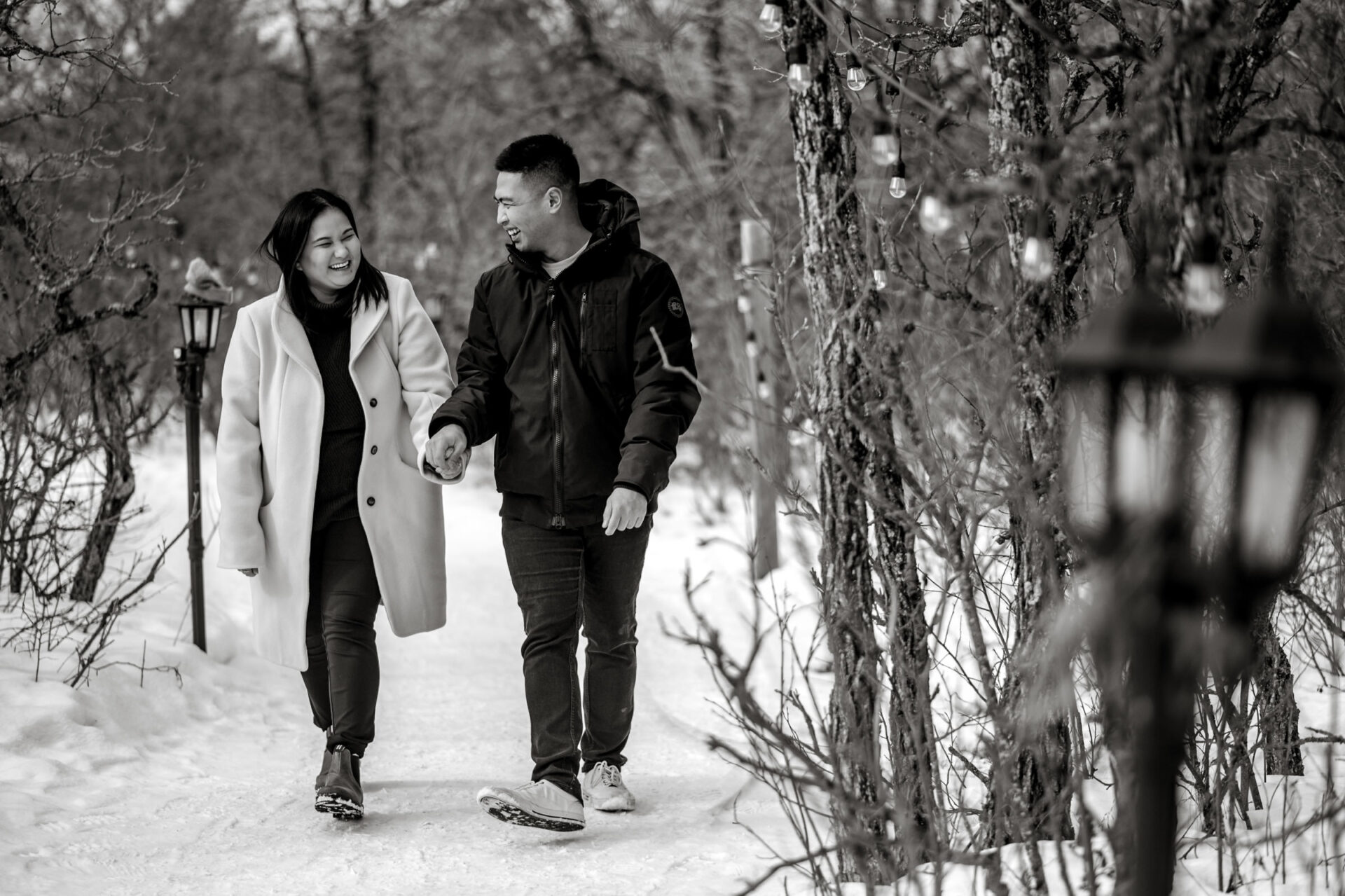 A couple enjoys a winter engagement on a snowy path lined with trees and lanterns.