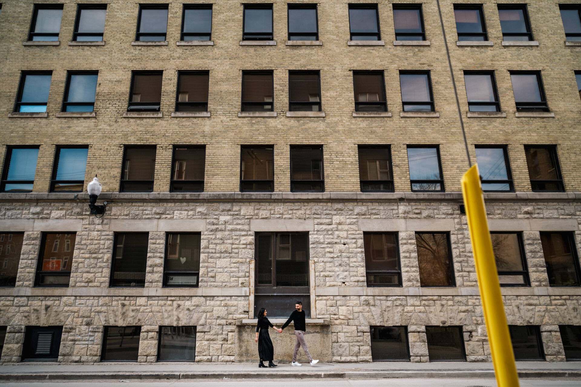 A couple holds hands on a sidewalk in Downtown Winnipeg, by a large brick building.