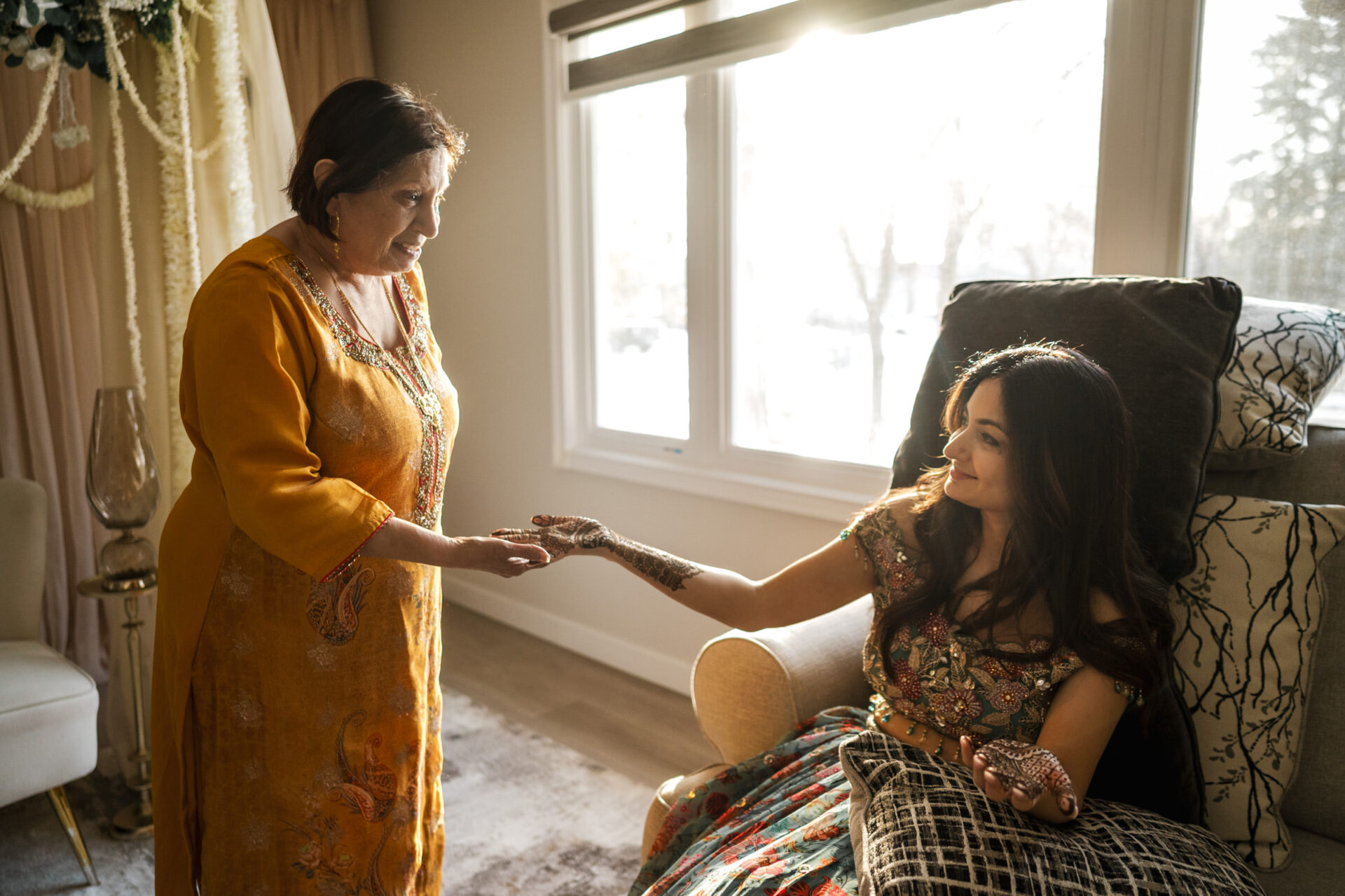 Woman in traditional attire helps another with henna at a warmly lit Hindu wedding.