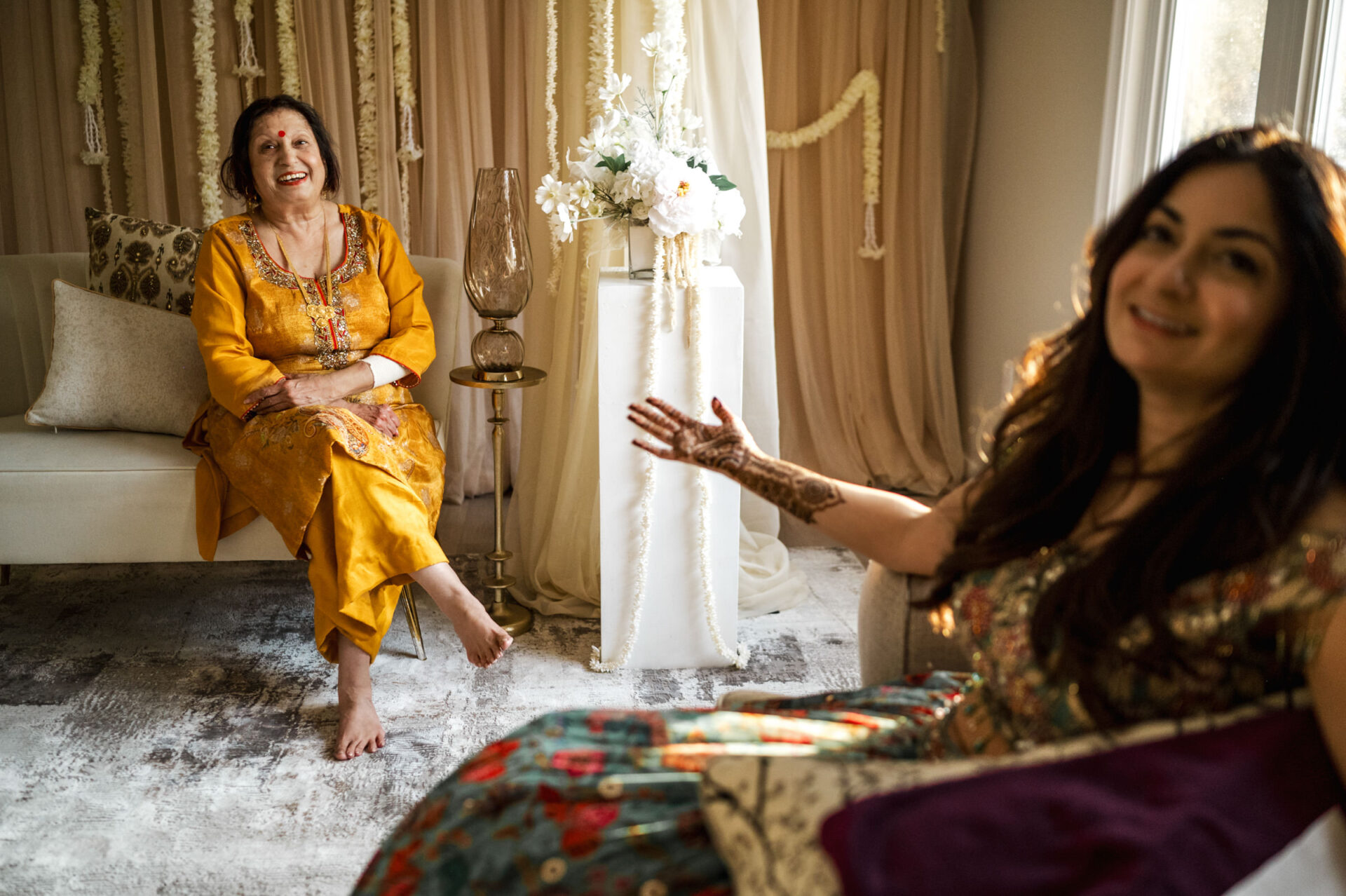 Two women smiling and chatting at a warmly lit Sikh wedding with elegant decor.
