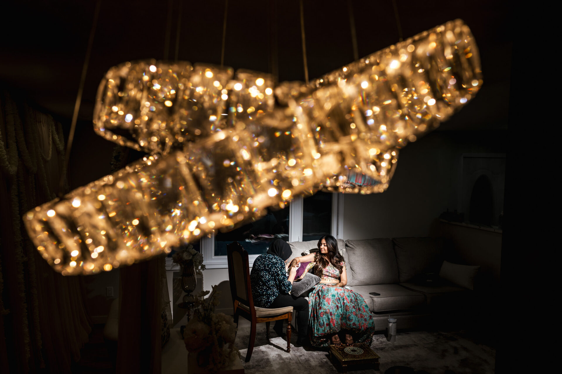 Elegant chandelier over two people discussing a Sikh wedding on a sofa in a dimly lit room.