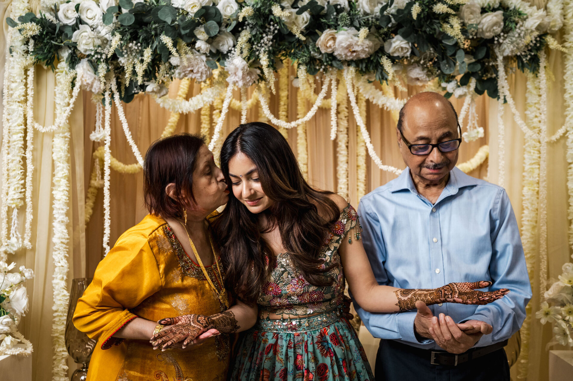 A woman with henna is embraced by her parents under floral decorations at a Hindu wedding.