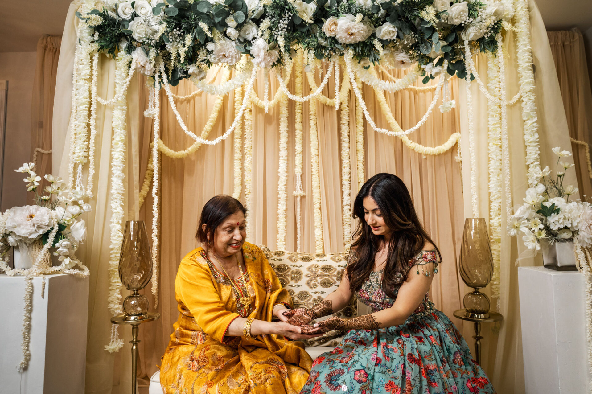 Two women in traditional attire share a moment under a flower-laden Sikh wedding canopy.