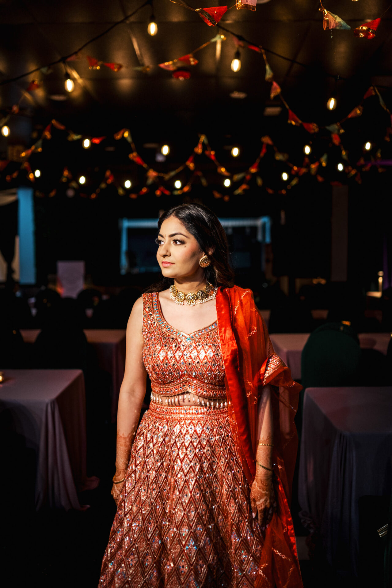 Woman in a red outfit at a Sikh wedding with string lights and draped tables.
