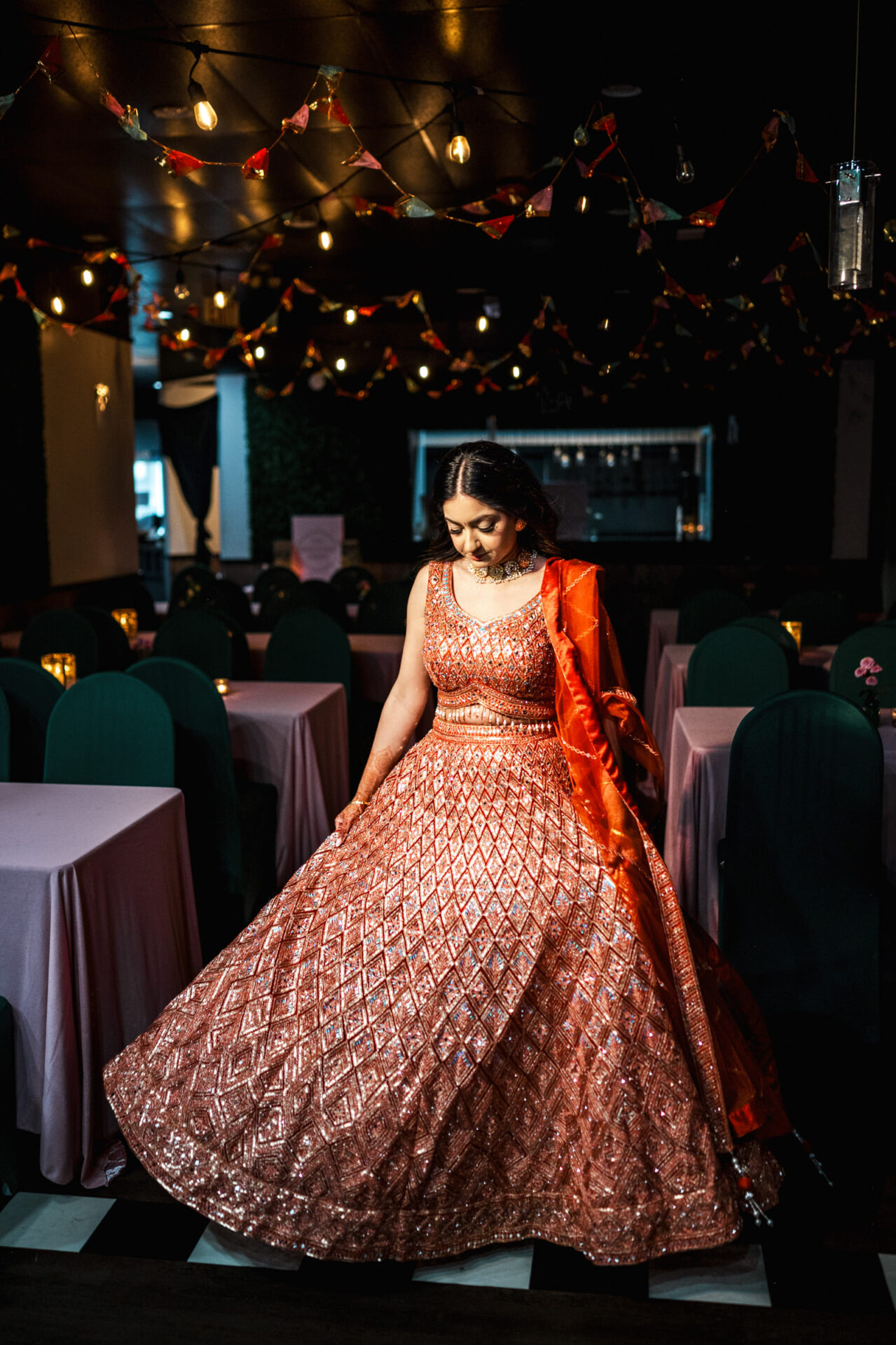 A woman twirls in a glittering pink lehenga at a dimly lit Sikh wedding.