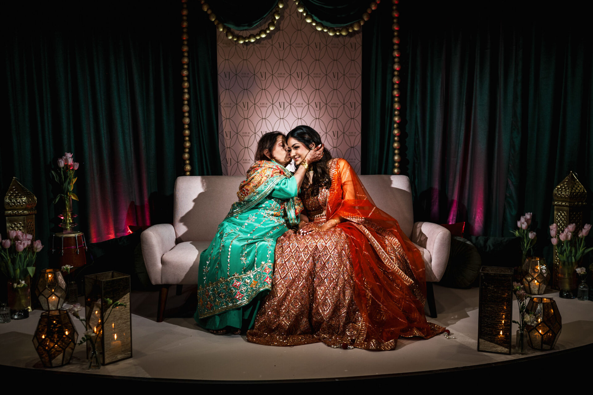 Two women in vibrant dresses share a joyful moment on a lantern-lit Sikh wedding stage.