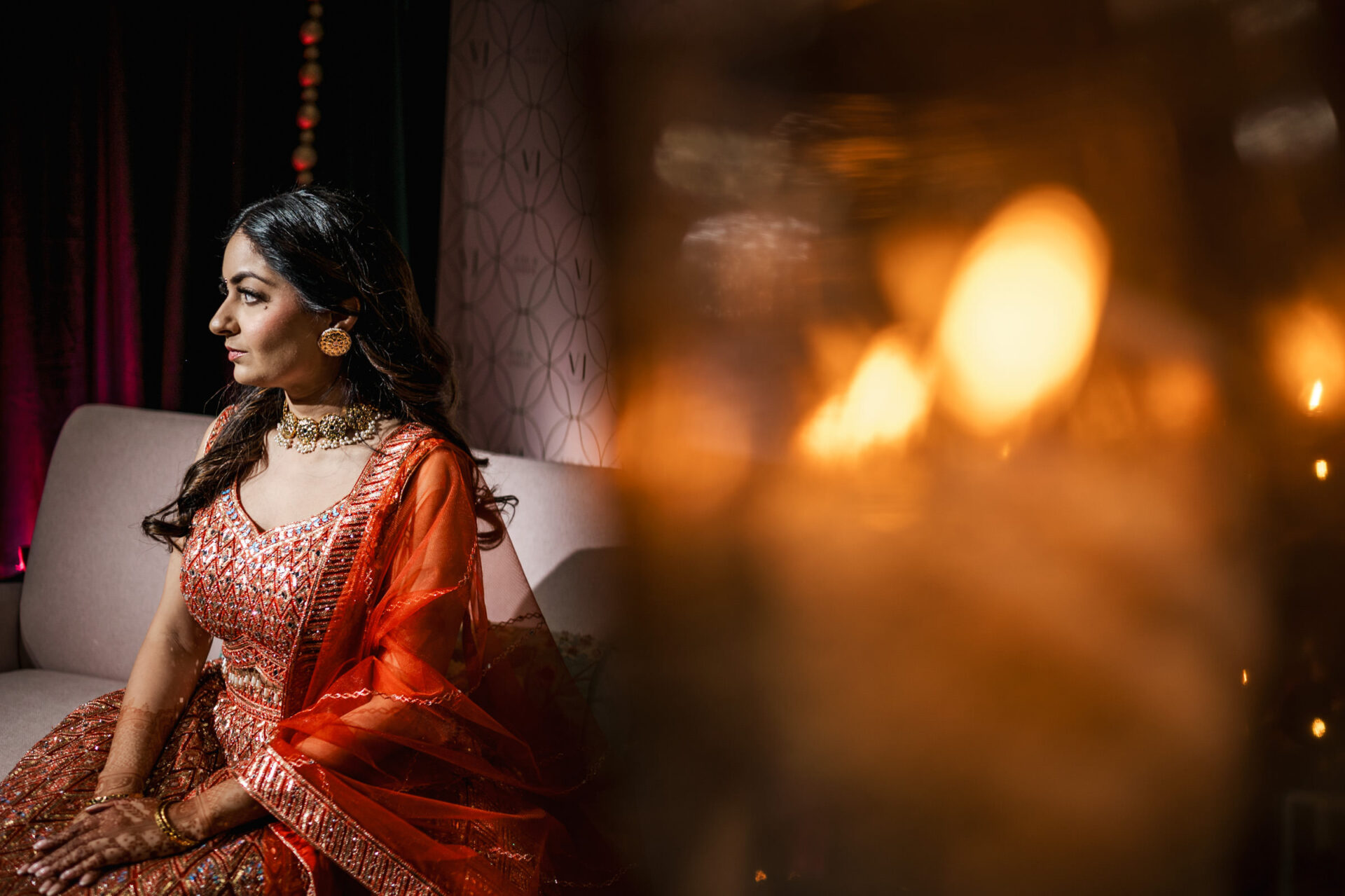 Woman in ornate dress sits on couch, warm lighting, looking left, festive wedding backdrop.
