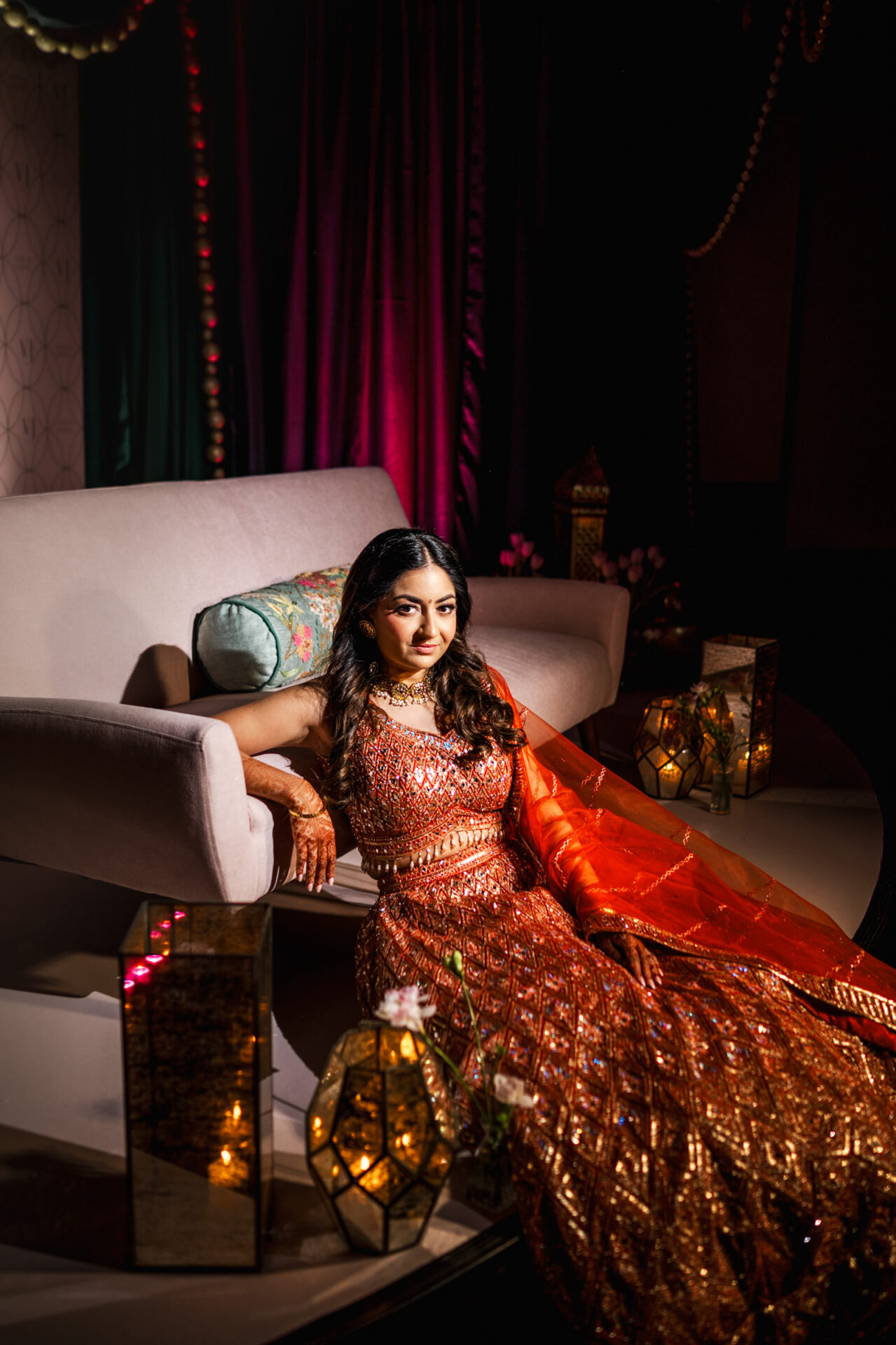 Woman in ornate orange dress at a Sikh wedding, surrounded by glowing lanterns and draped fabrics.