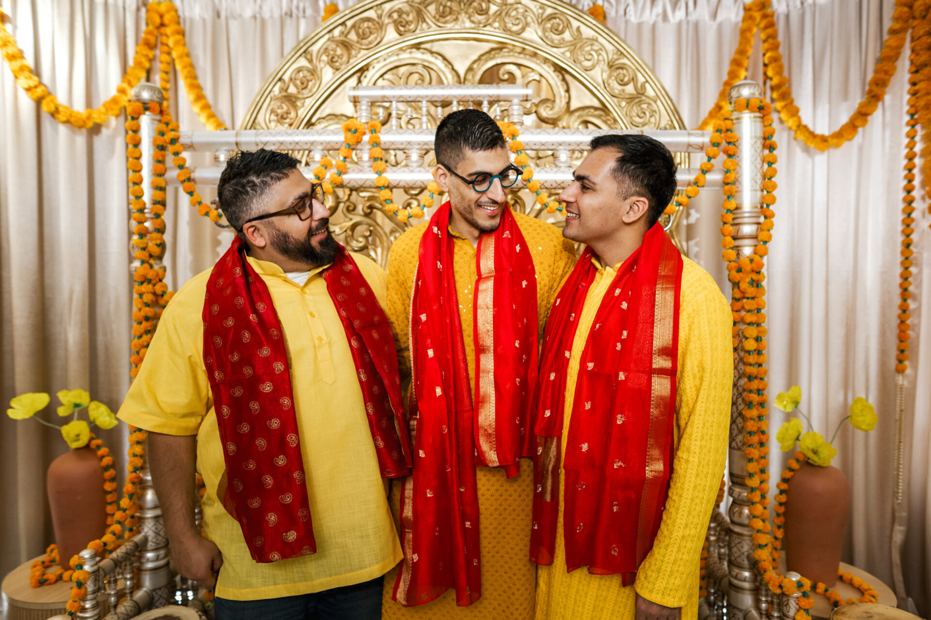 Three men in yellow and red smile at a vibrant Sikh wedding celebration indoors.