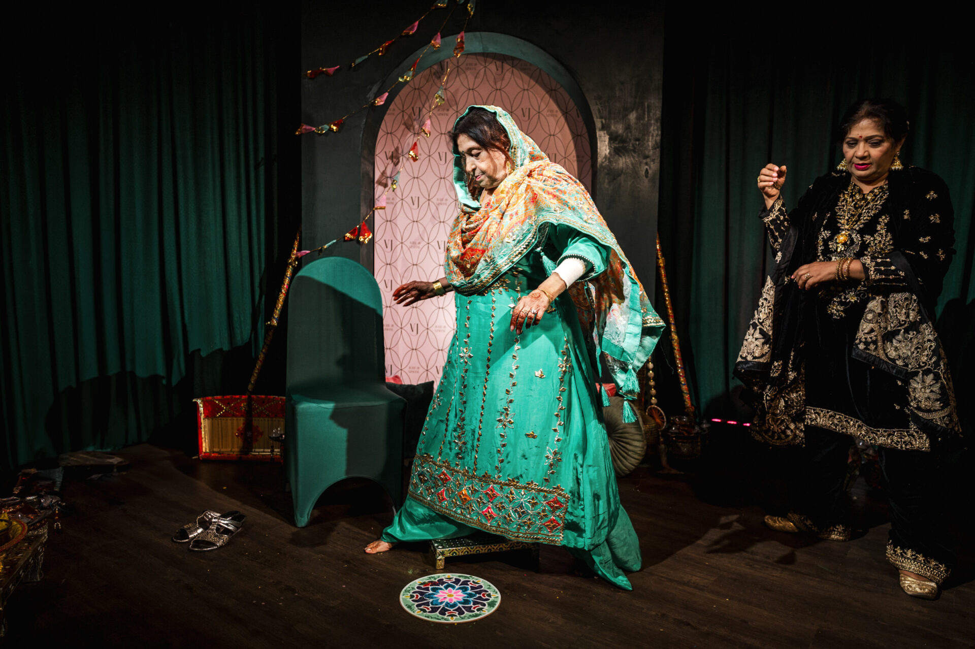 Two women in colorful attire perform at a Sikh wedding with a decorative backdrop.
