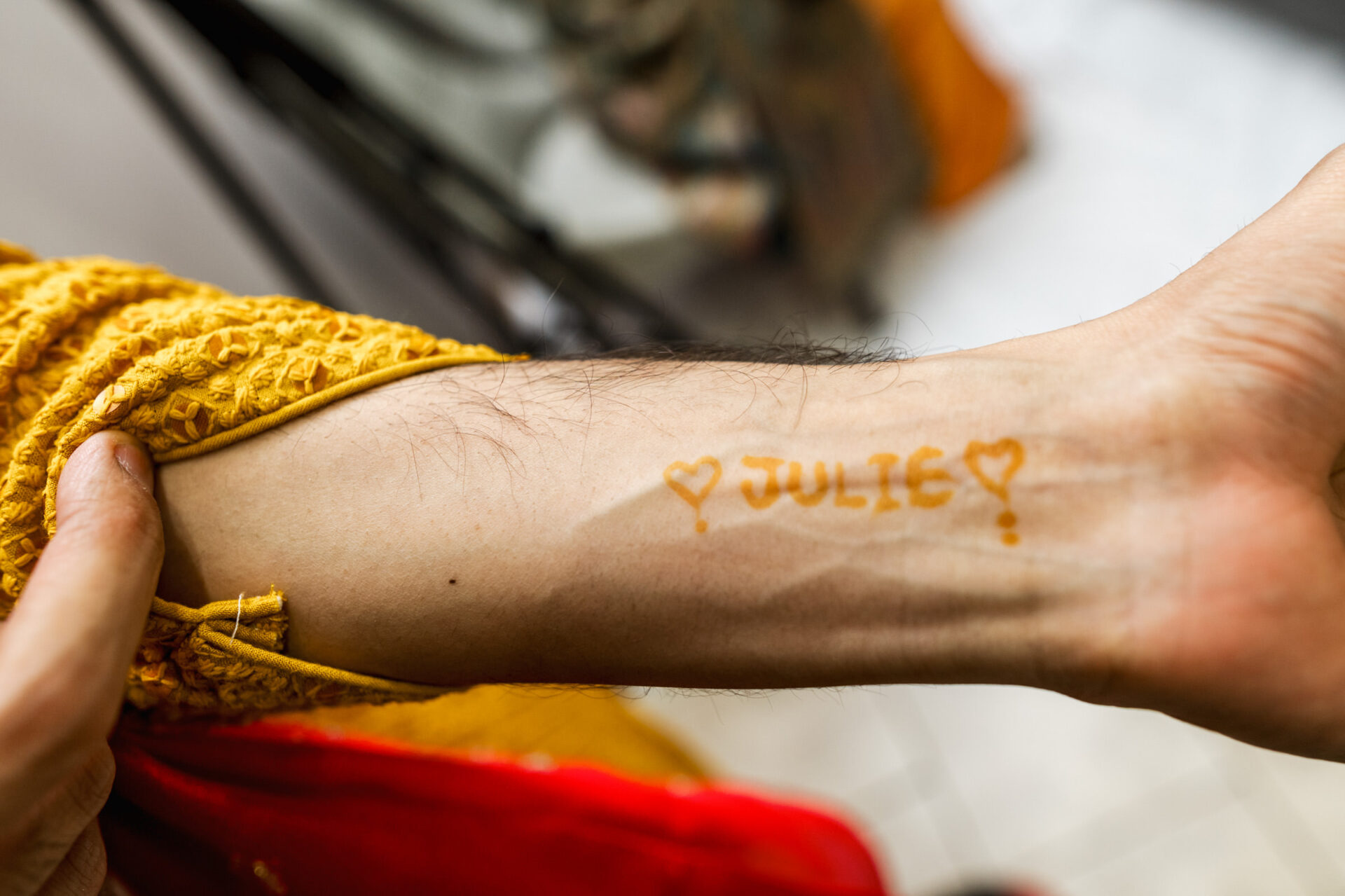 Arm with "JULIE" and hearts on skin, wearing yellow textured fabric at a Sikh wedding.