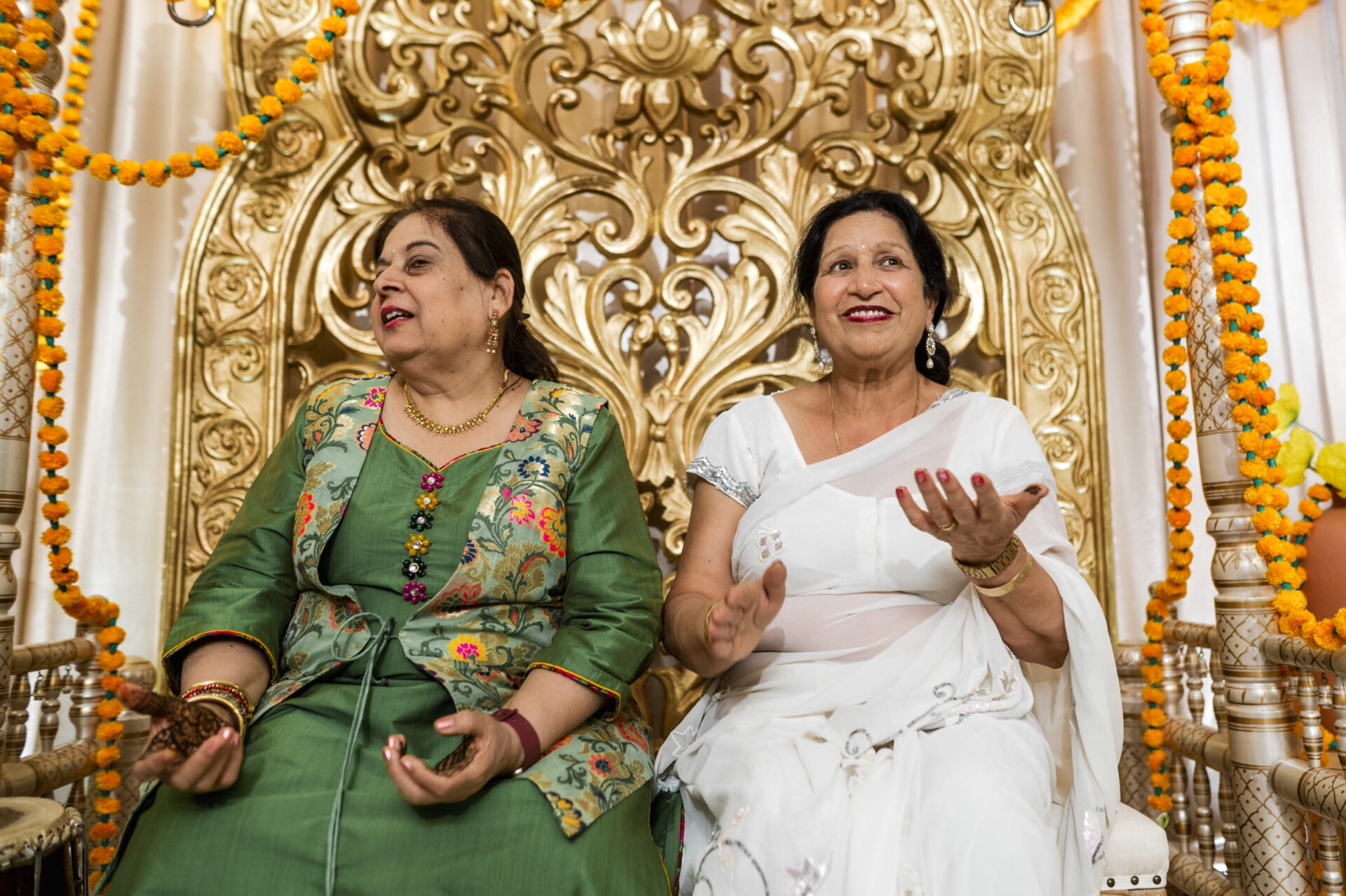 Two women in colorful attire, smiling and clapping at a vibrant Sikh wedding celebration.