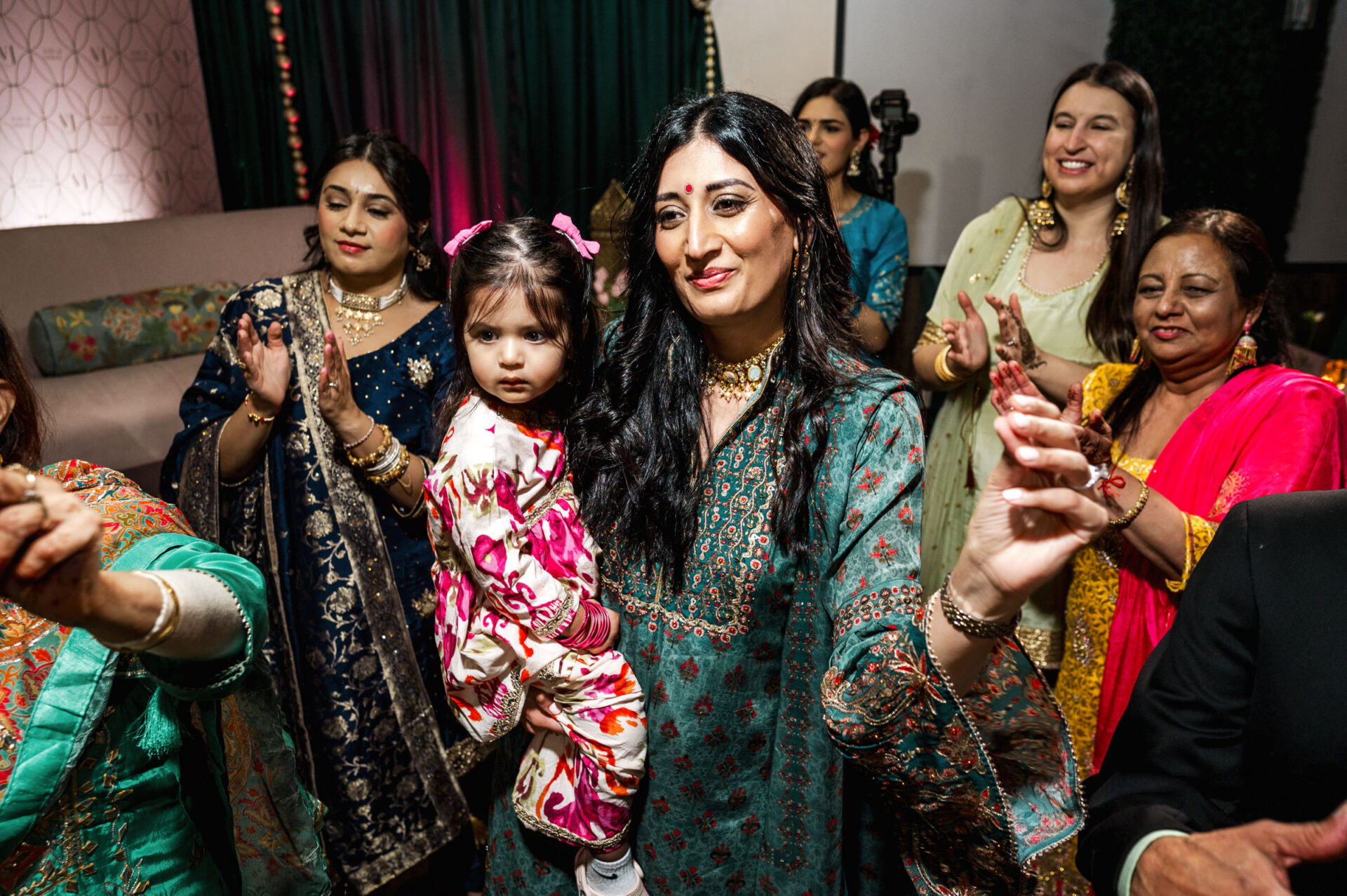 A group celebrates a vibrant Sikh wedding; woman holds child as others clap.
