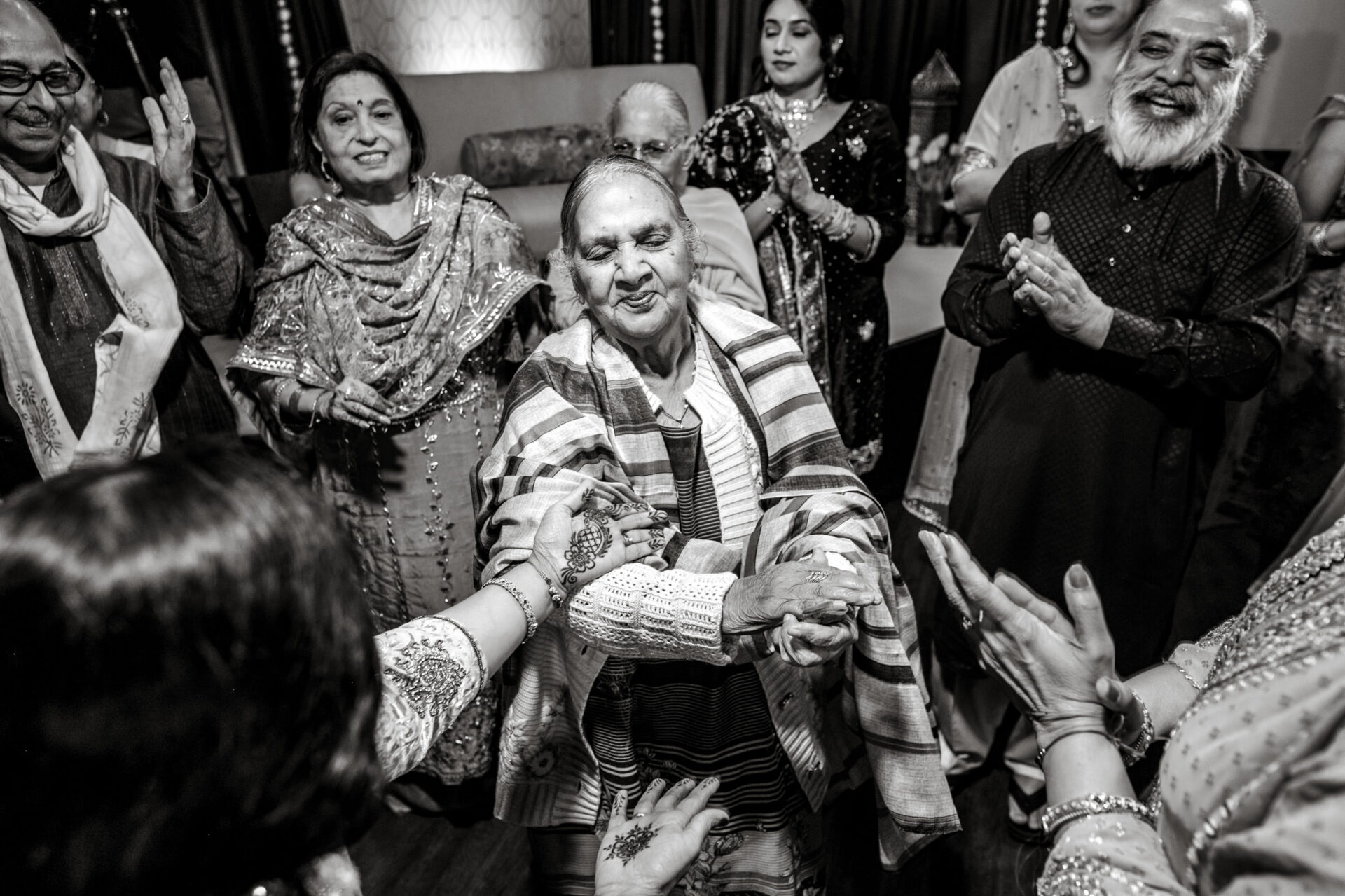 A joyful elderly woman dances at a lively Sikh gathering, surrounded by clapping people.