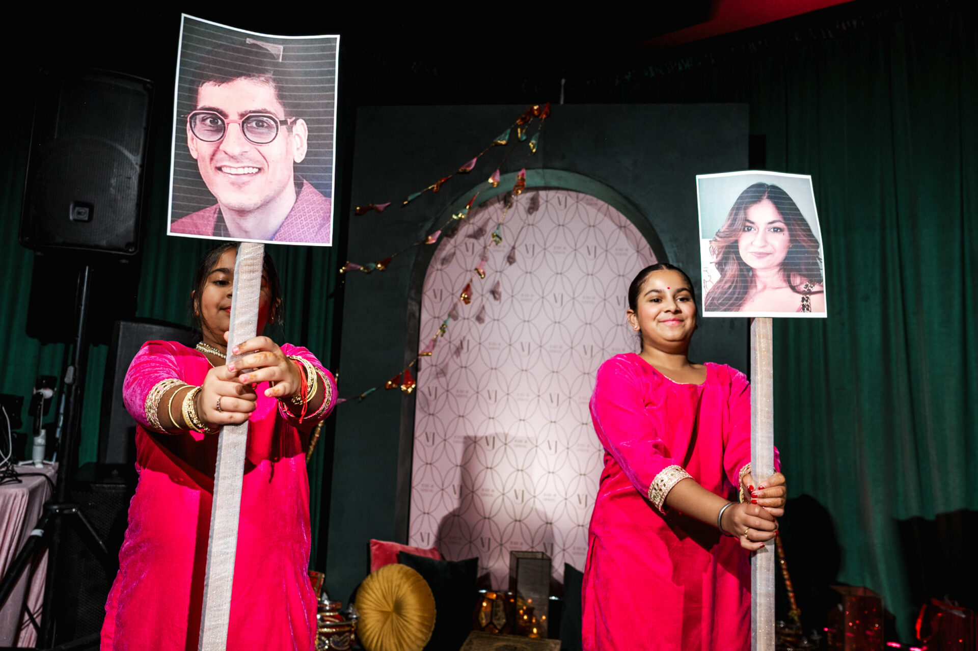 Two people in pink outfits hold photos on sticks at a vibrant Sikh wedding event.