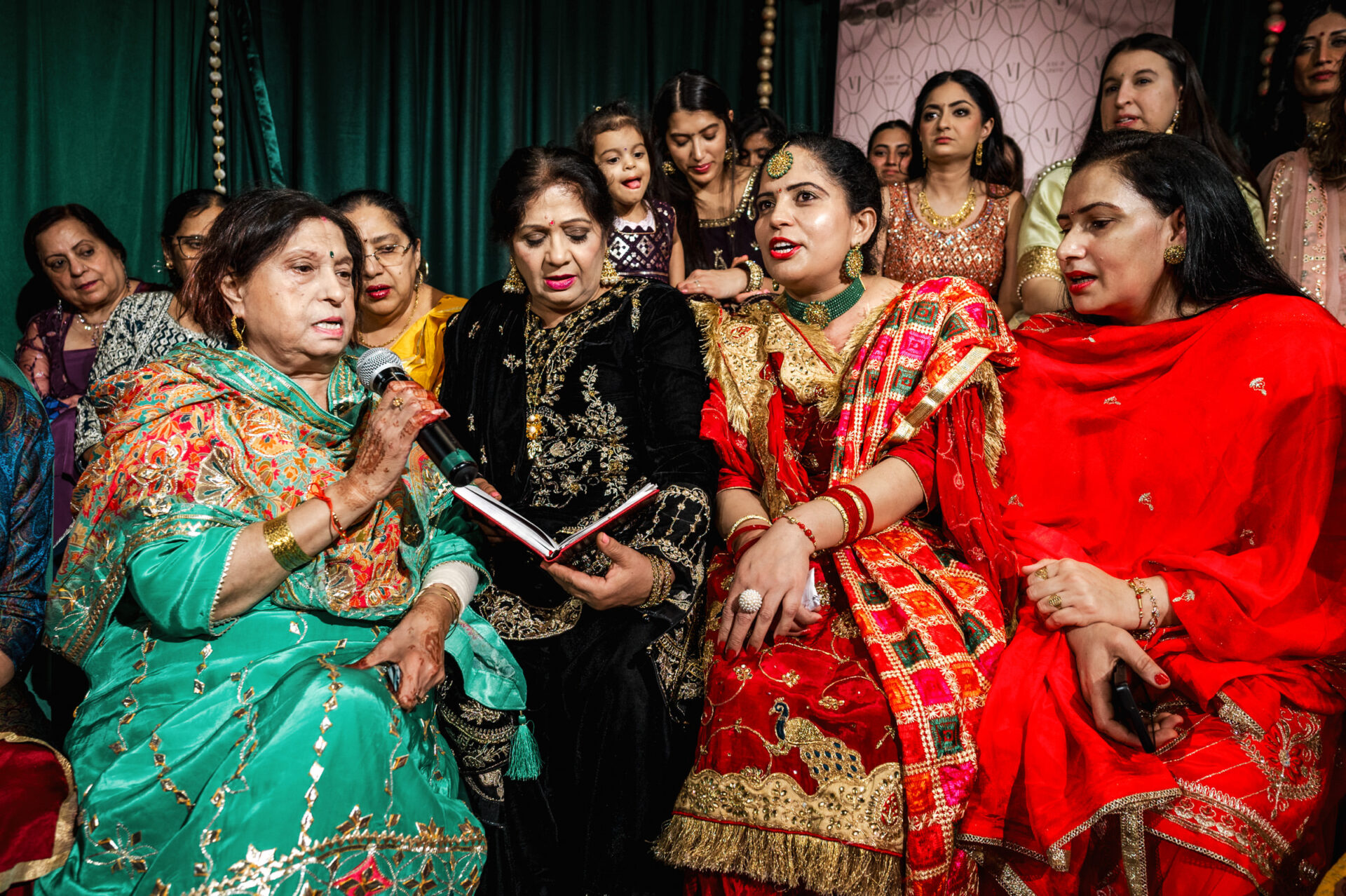 A group of women in vibrant traditional clothing sing together at a lively Sikh wedding.