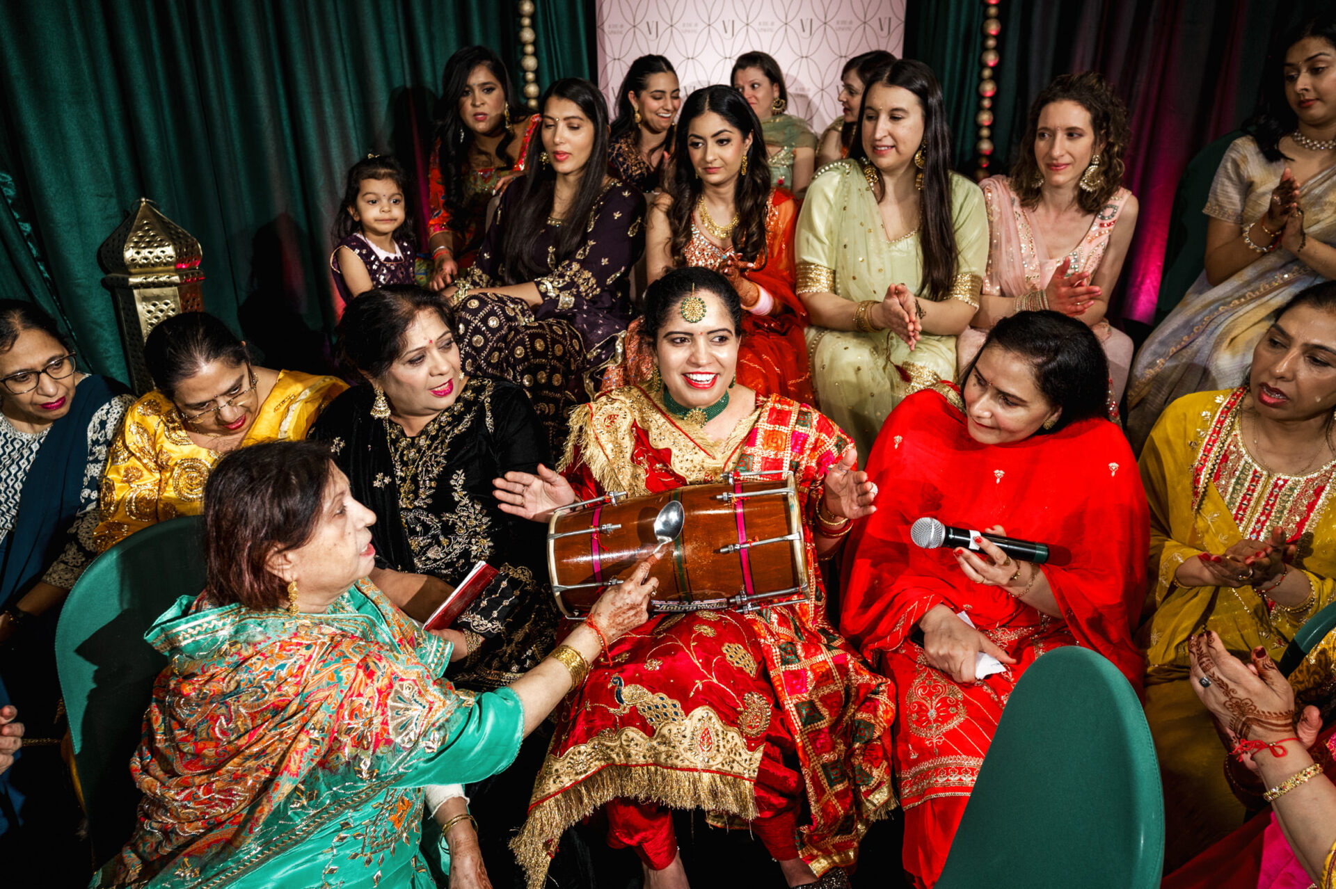 Women and girls at a lively Sikh event, one playing a dholak, in colorful attire.