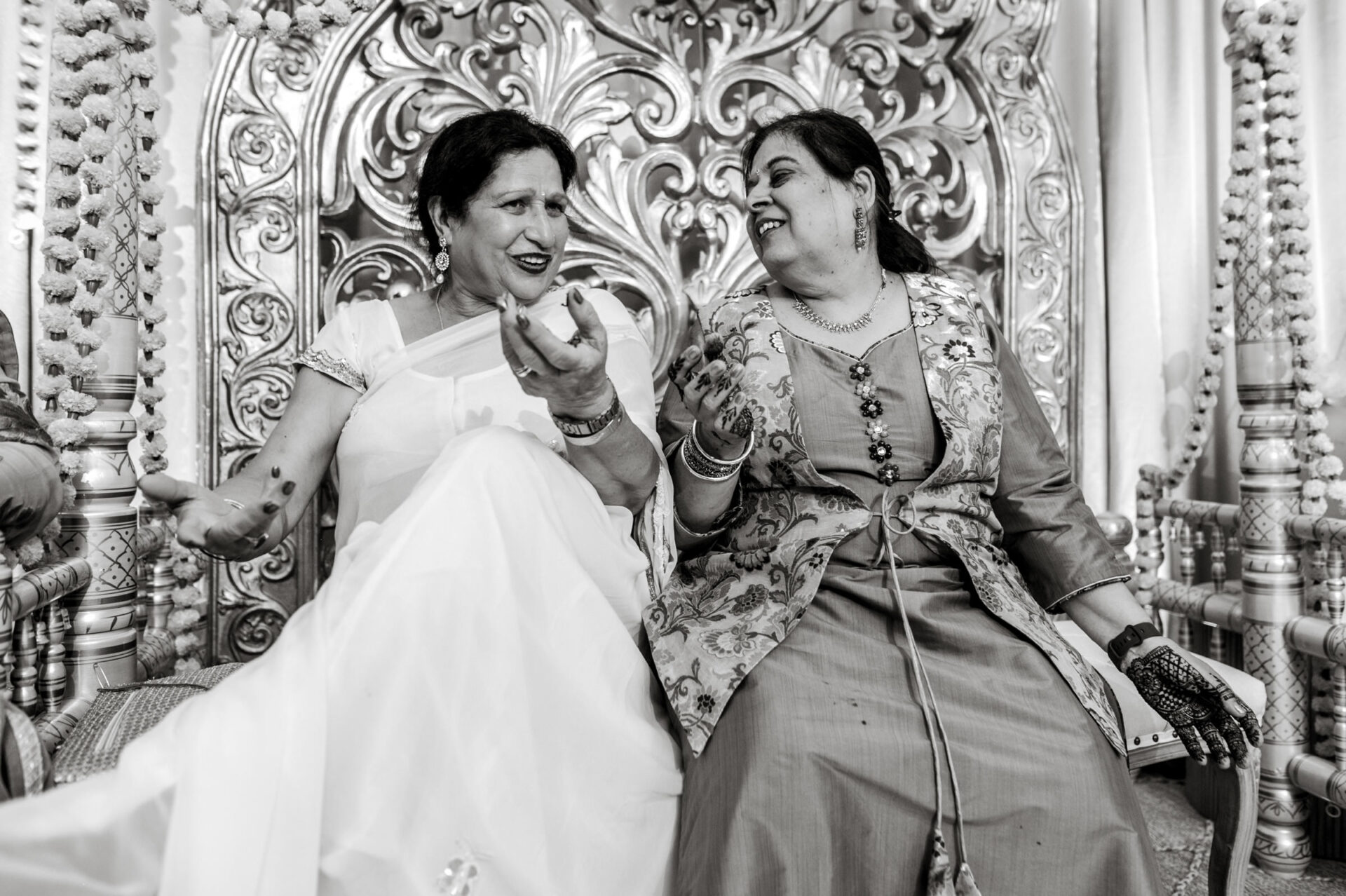 Two women laughing, sitting on a decorated chair at a vibrant Sikh wedding.