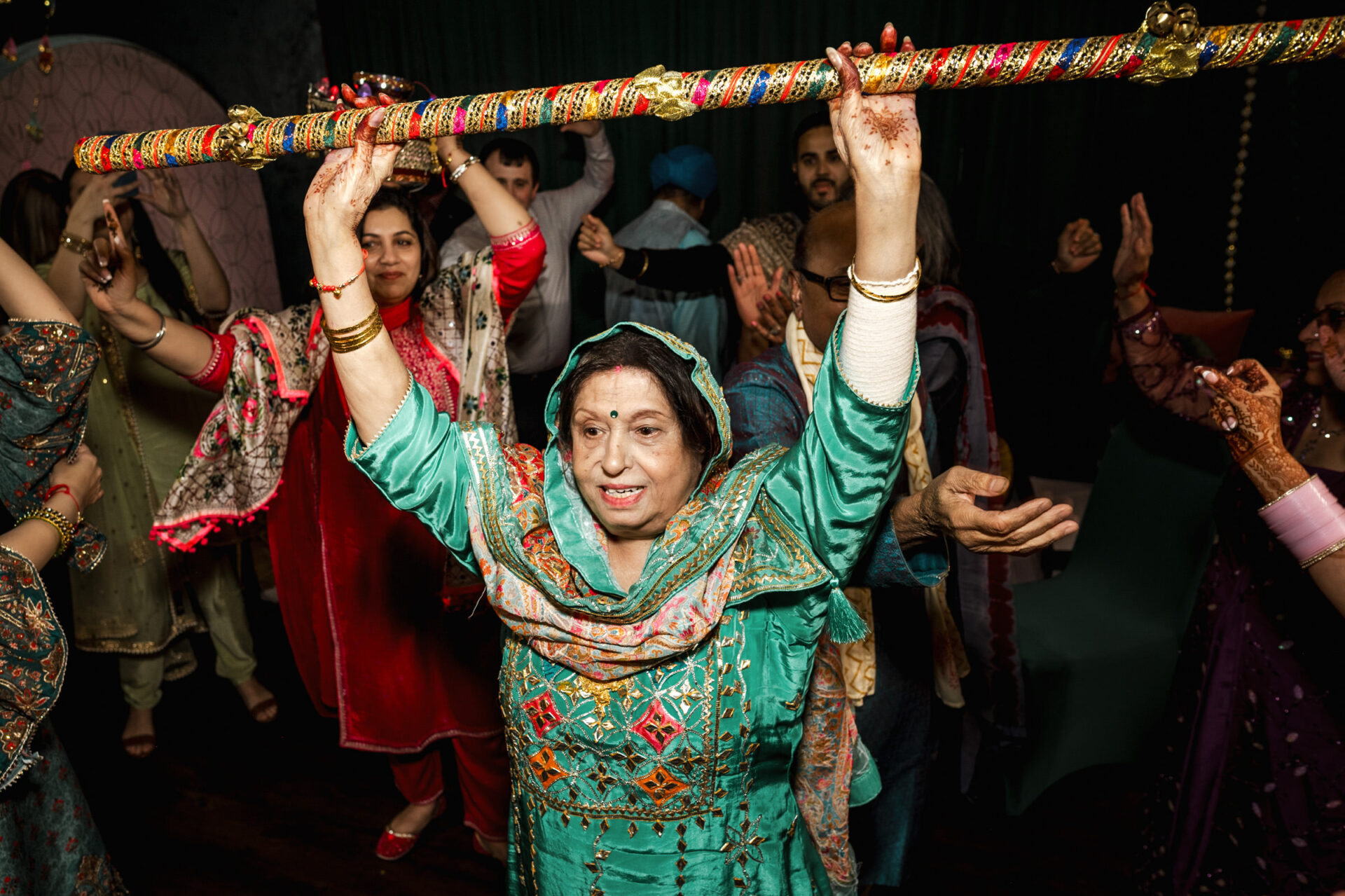 Woman in traditional attire dances joyfully, stick overhead at a Sikh wedding.
