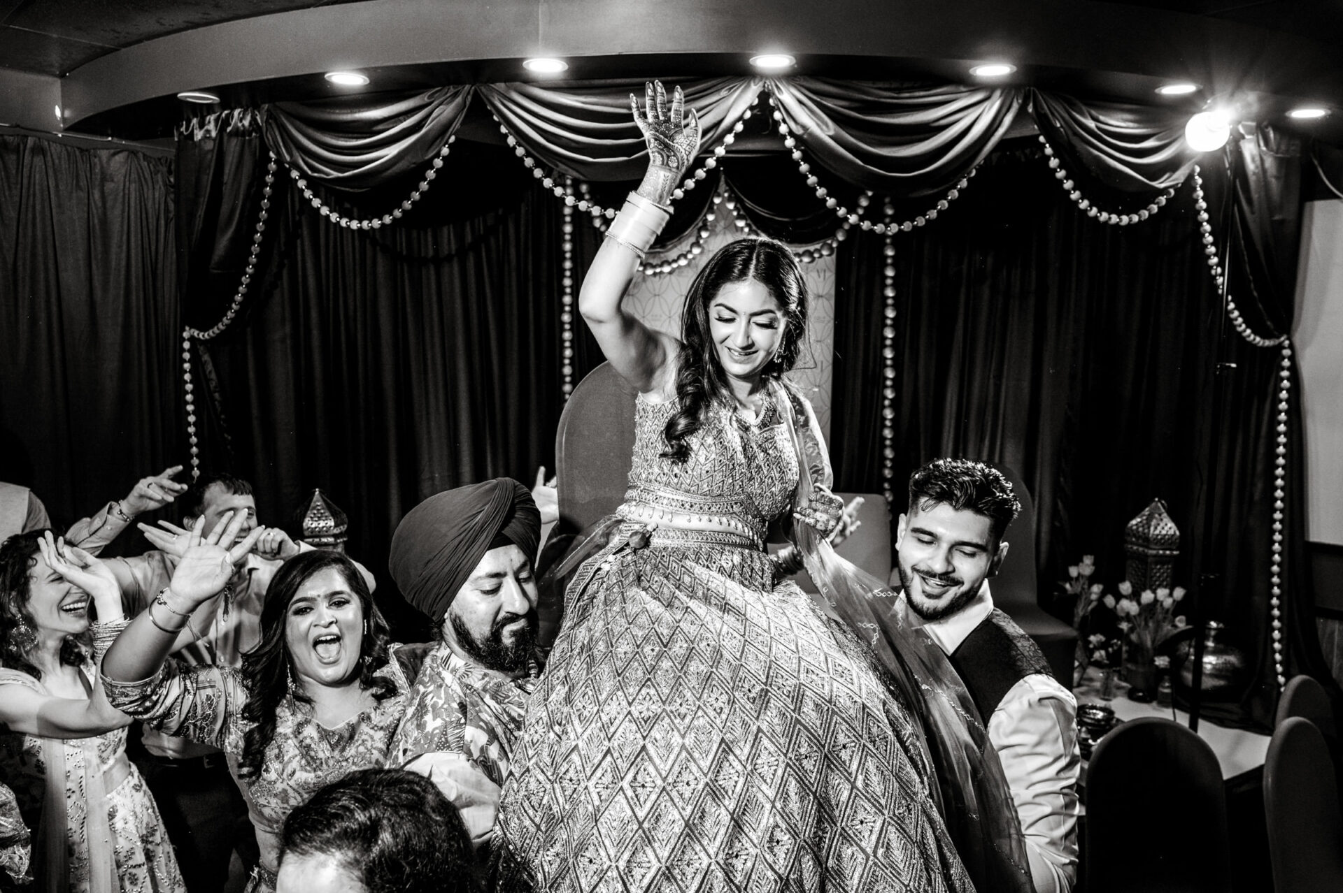 Bride joyfully lifted on chair at lively indoor Sikh wedding celebration.