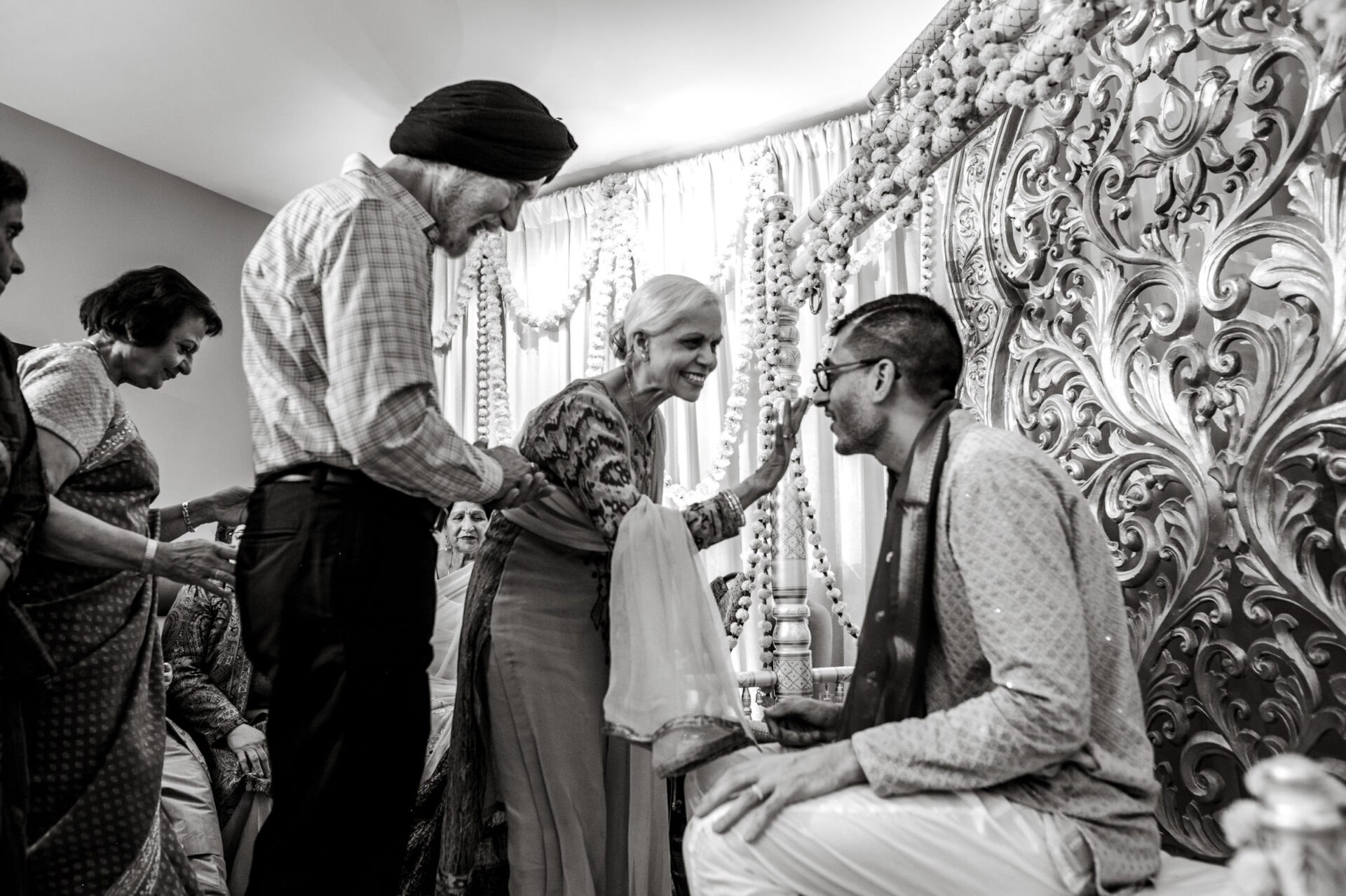Elderly couple greeting seated man at a vibrant Sikh wedding, surrounded by others.