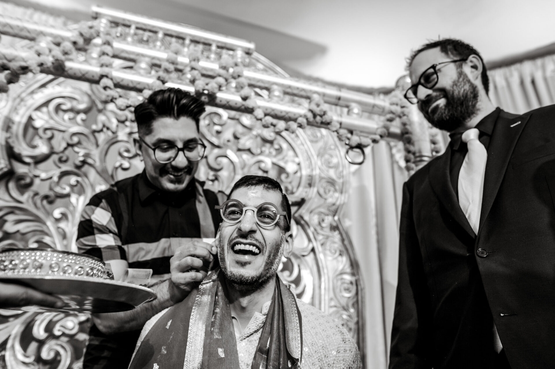 Three men at a Sikh celebration, one laughing, adorned in traditional attire and intricate backdrop.