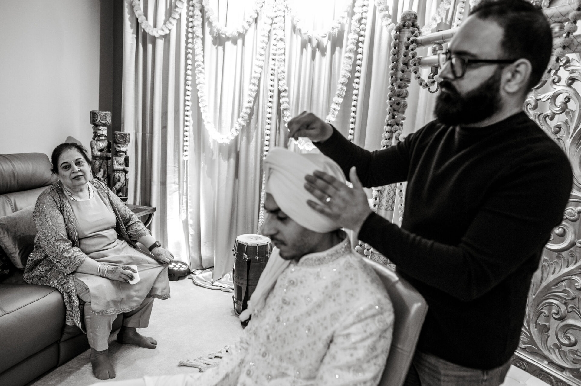 Sikh man helps groom with turban as a woman sits nearby in a decorated room.