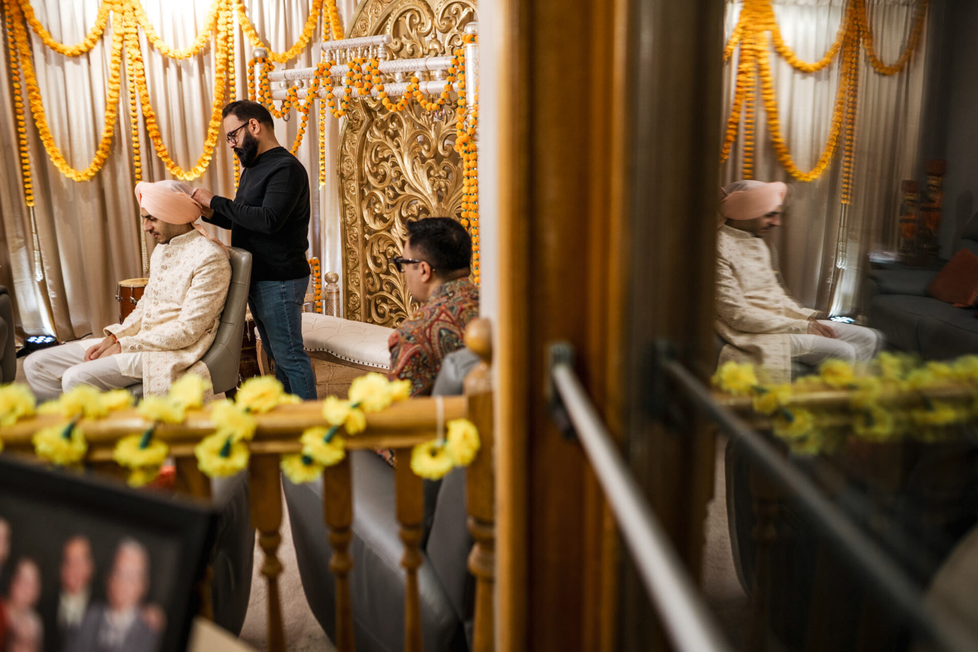 Man adjusting turban of another at a vibrant Sikh wedding in a garland-adorned room.