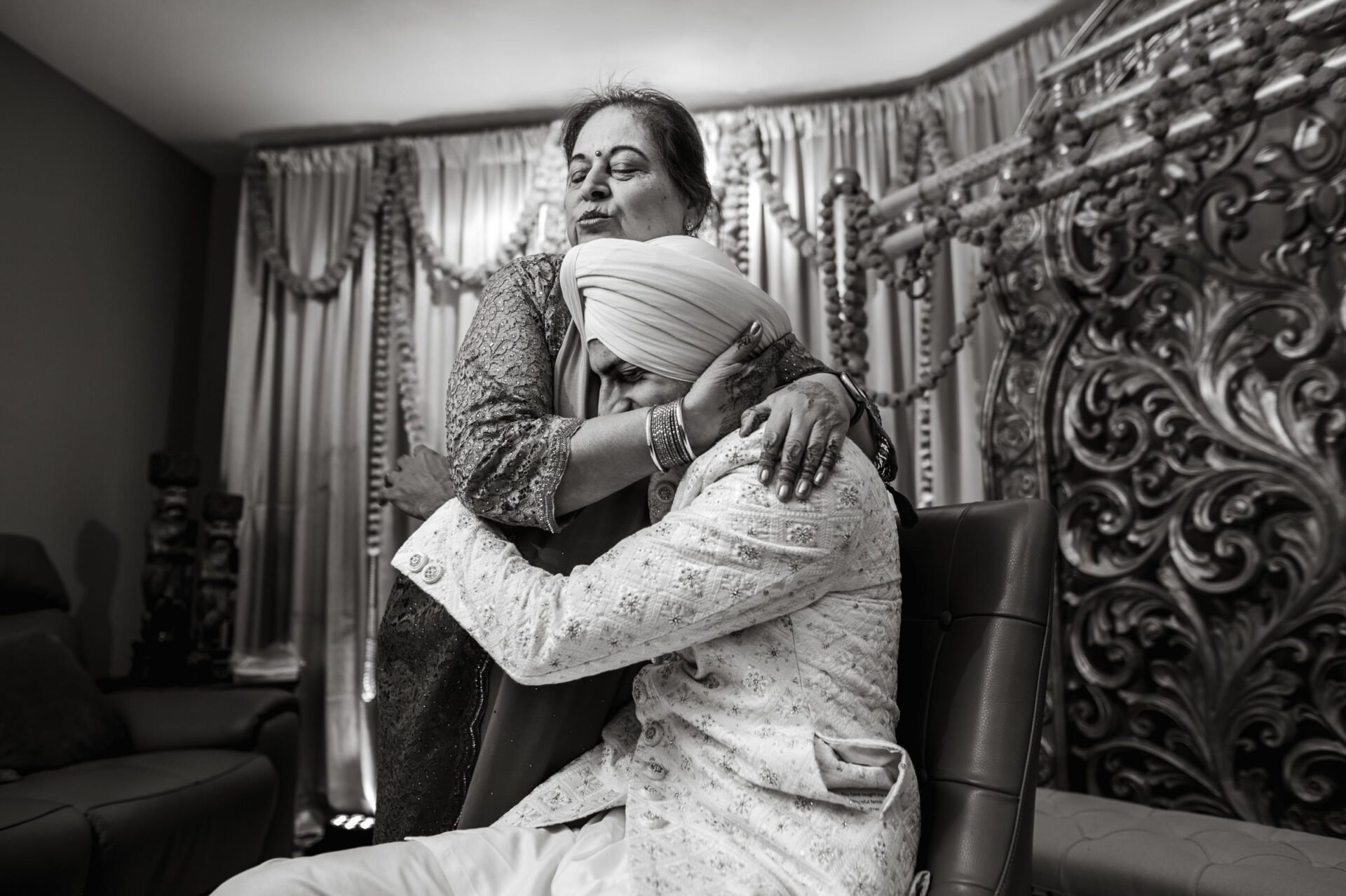 A joyful embrace at a Sikh wedding between a seated turbaned person and standing woman.