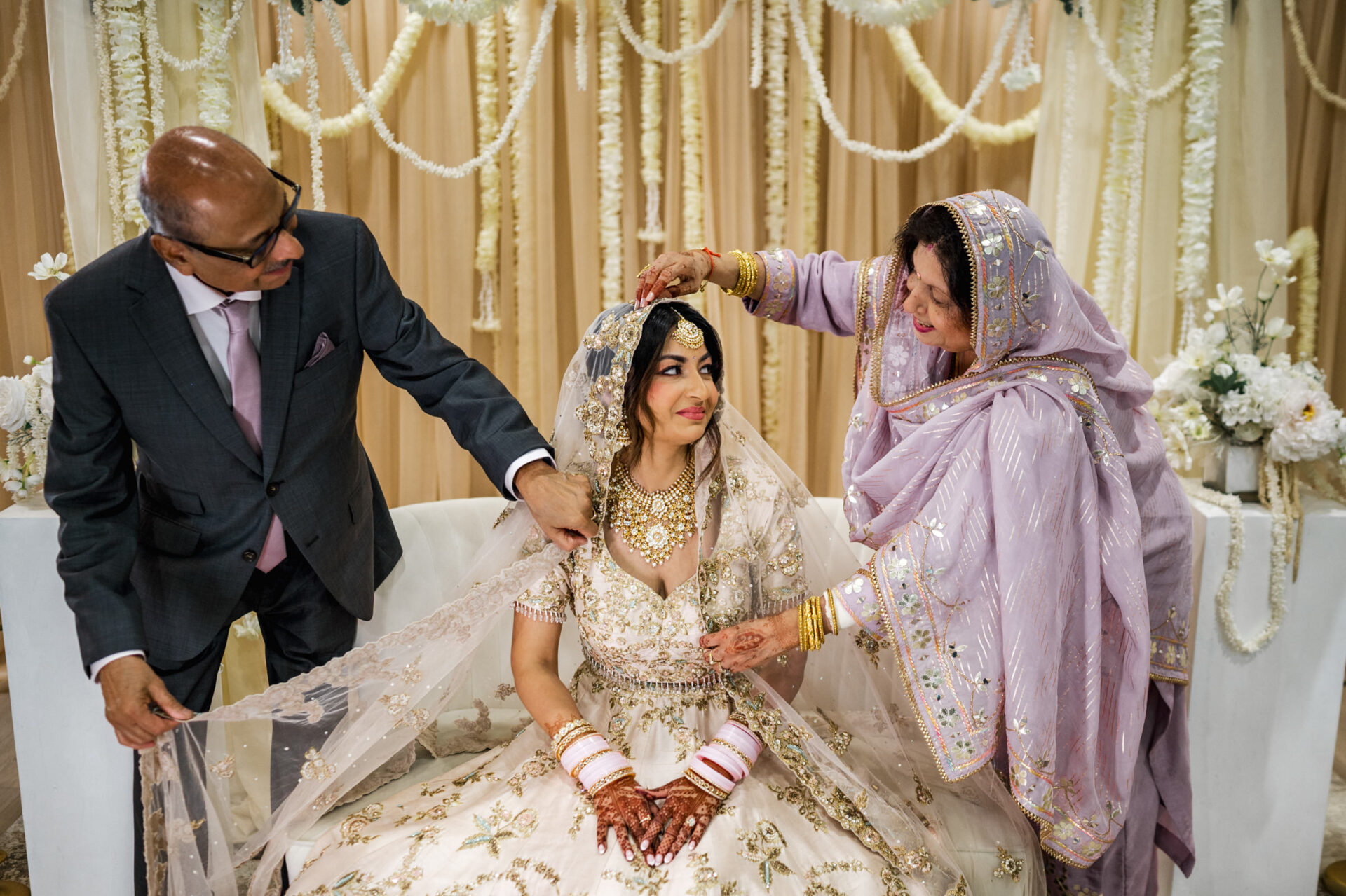 Bride in ornate dress at Sikh ceremony, parents adjusting her veil.