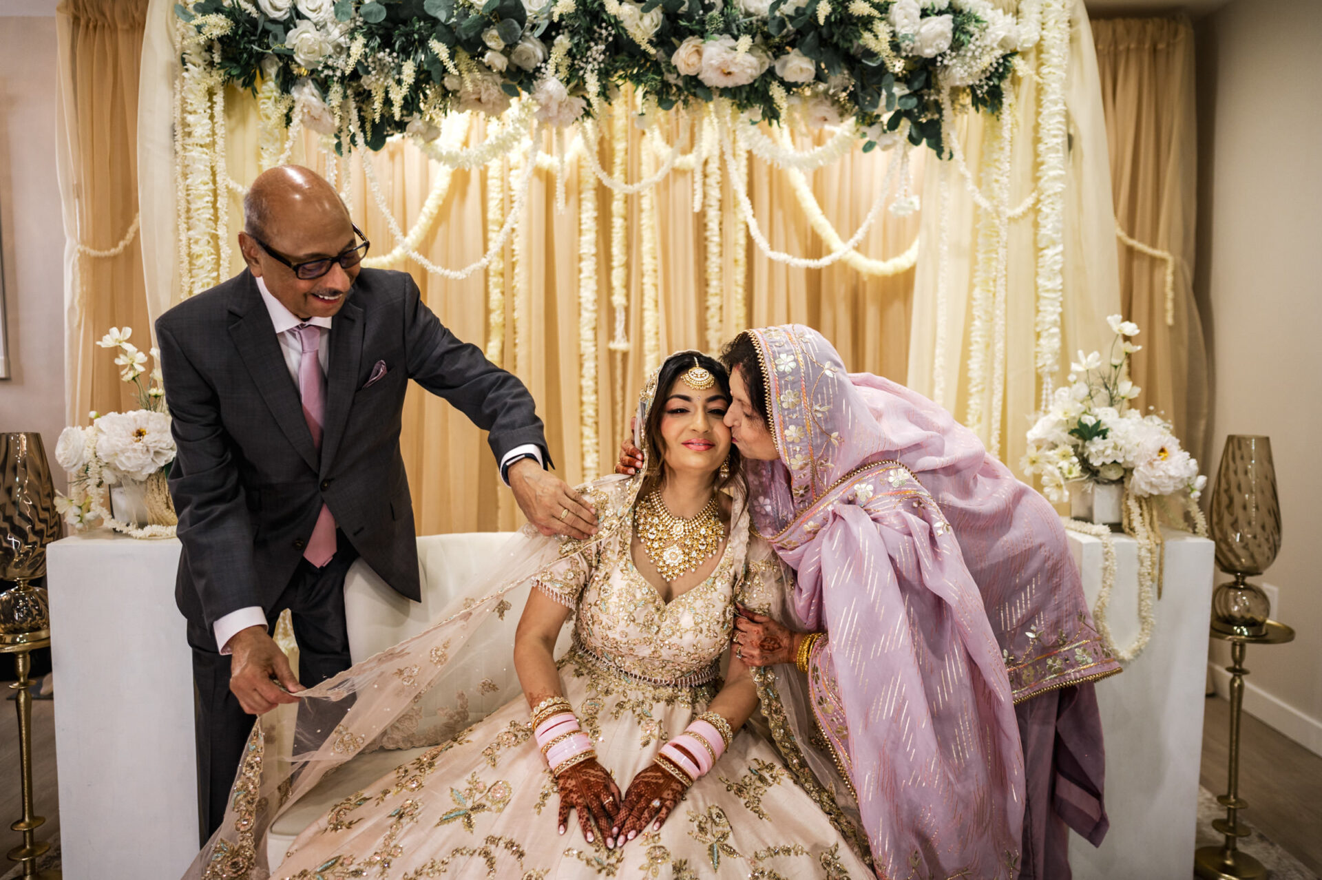 Bride in traditional attire kissed by a woman at Sikh wedding, man smiling beside them.