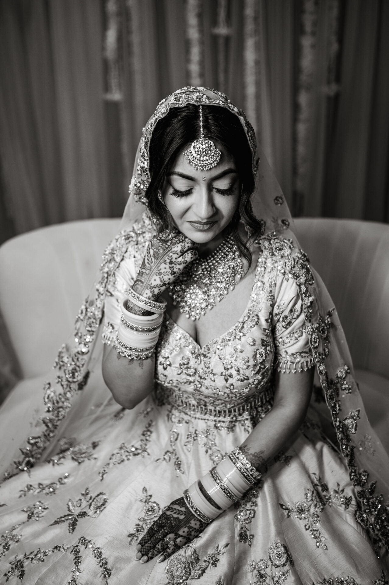 Bride in ornate outfit and jewelry, eyes closed, Henna on hands, at Sikh wedding in black and white.