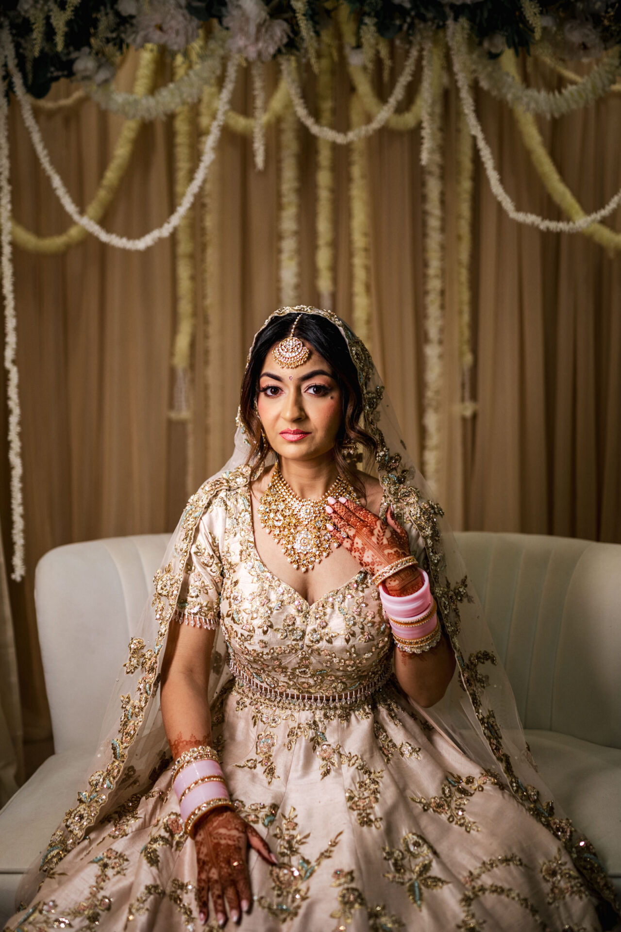Sikh bride in ornate dress and jewelry, seated, with floral decor in the background.