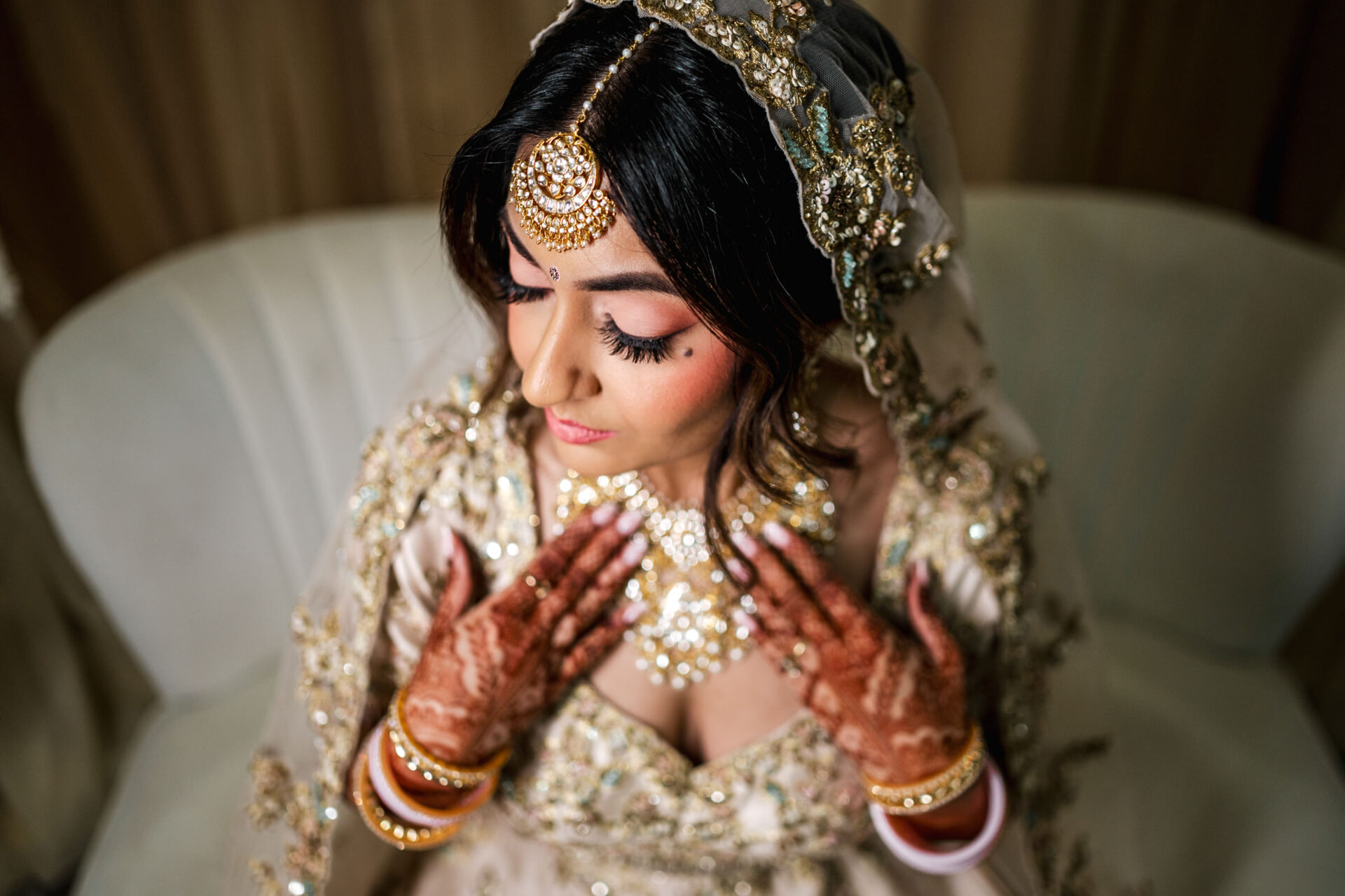 Sikh bride in ornate dress and jewelry, with henna, sits on a white couch.