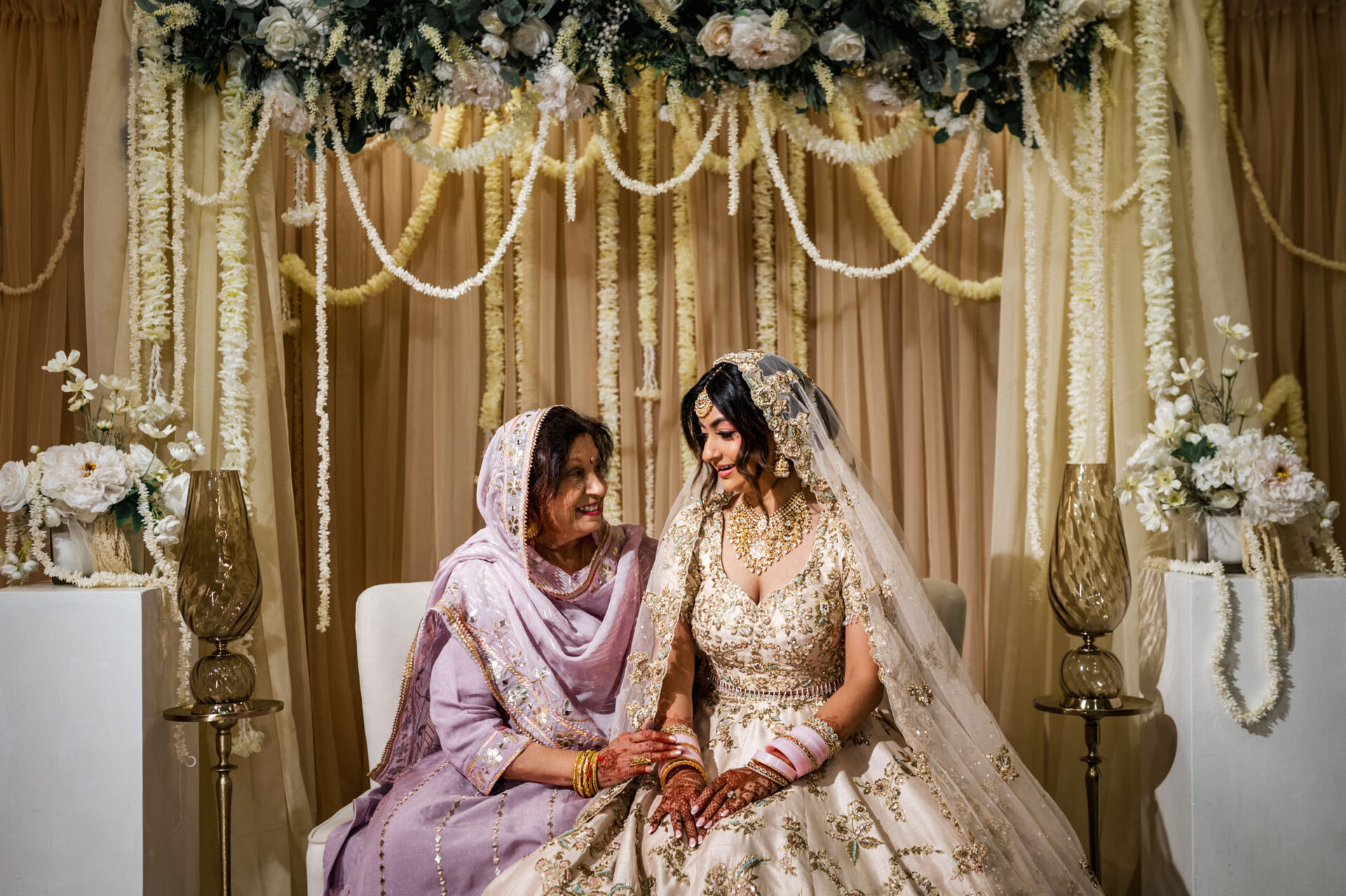 Bride and older woman smiling under a floral canopy at a Sikh wedding ceremony.