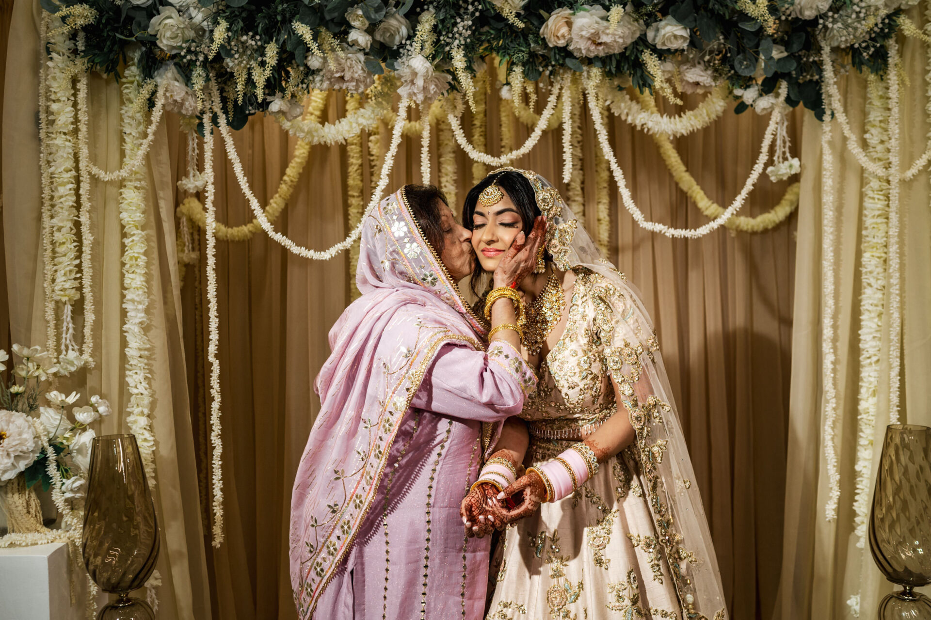 At a Sikh wedding, a woman in a sari kisses the bride's cheek under floral decorations.