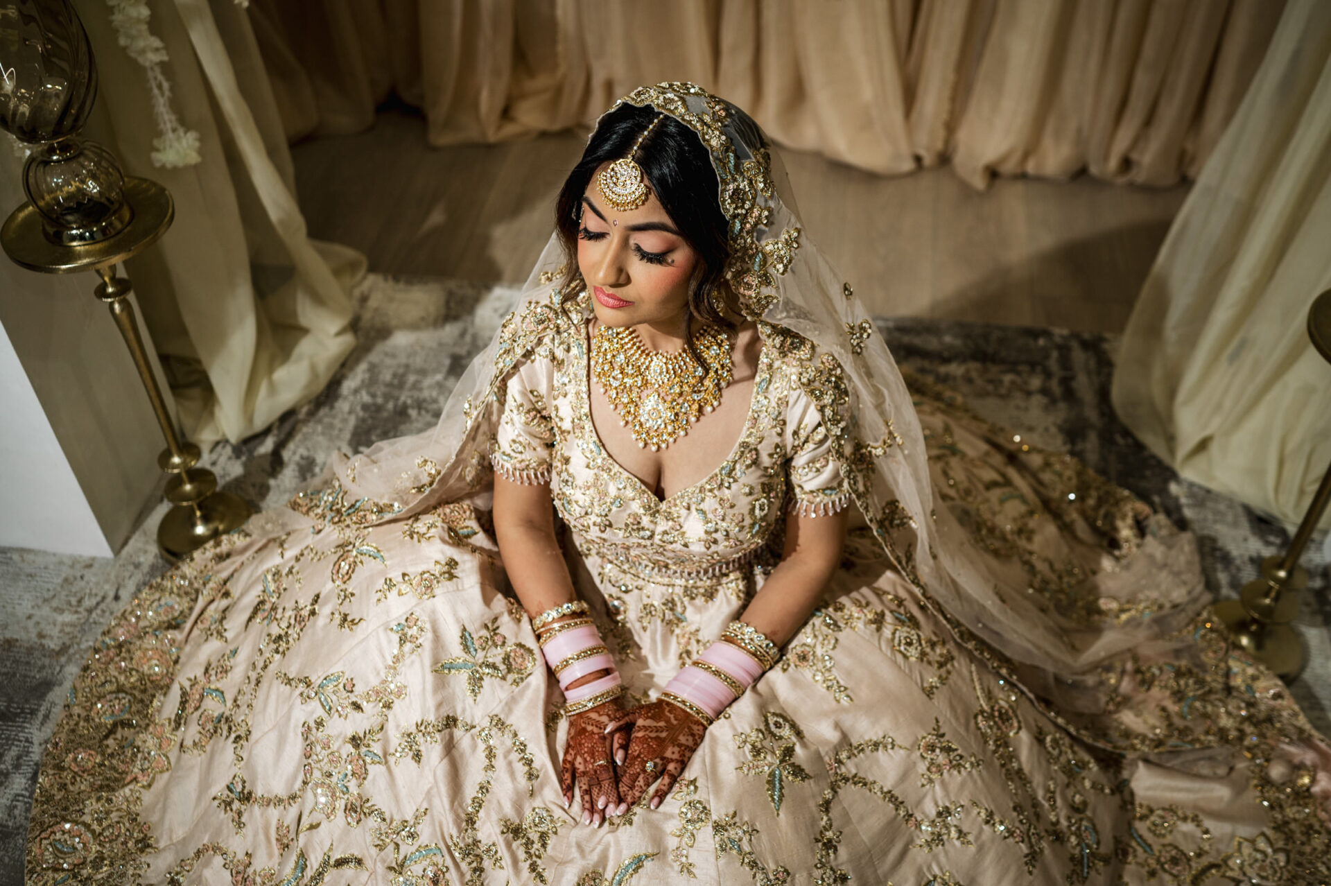 Bride in ornate cream lehenga with jewelry, sitting gracefully at a Sikh wedding on a rug.