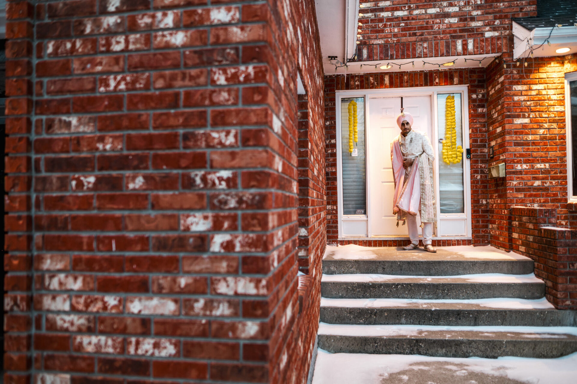 Person in traditional attire, like a Sikh groom, stands on snowy steps by the brick house.