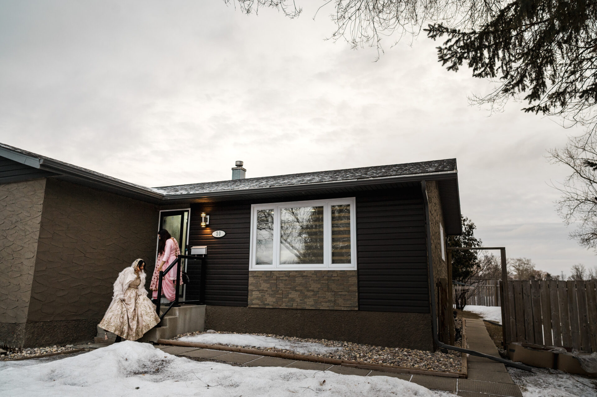 Two people in winter clothing enter a house, snow covering the ground like a wedding veil.