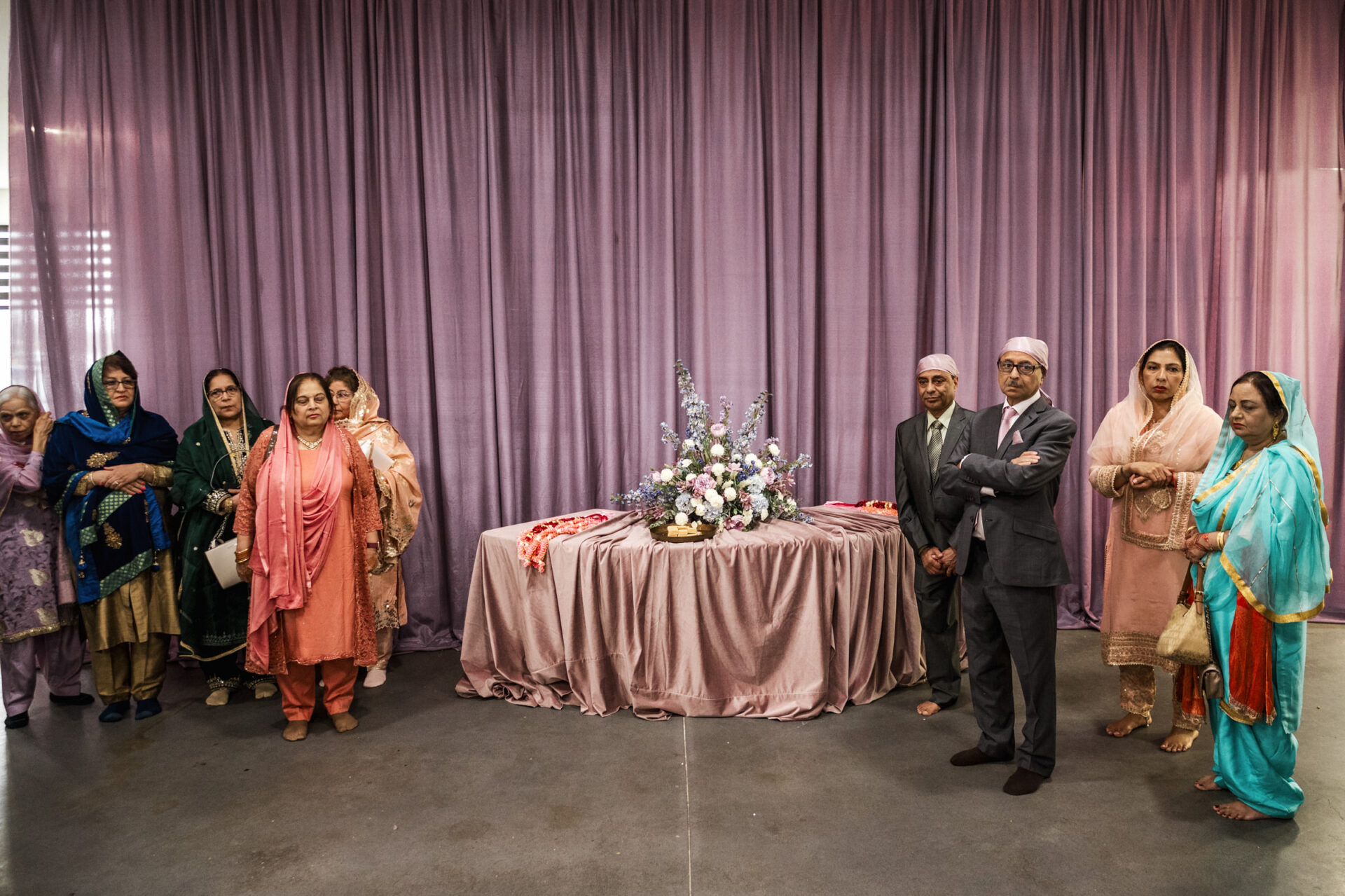 A Sikh gathering in traditional attire around a table with flowers, set against a purple backdrop.