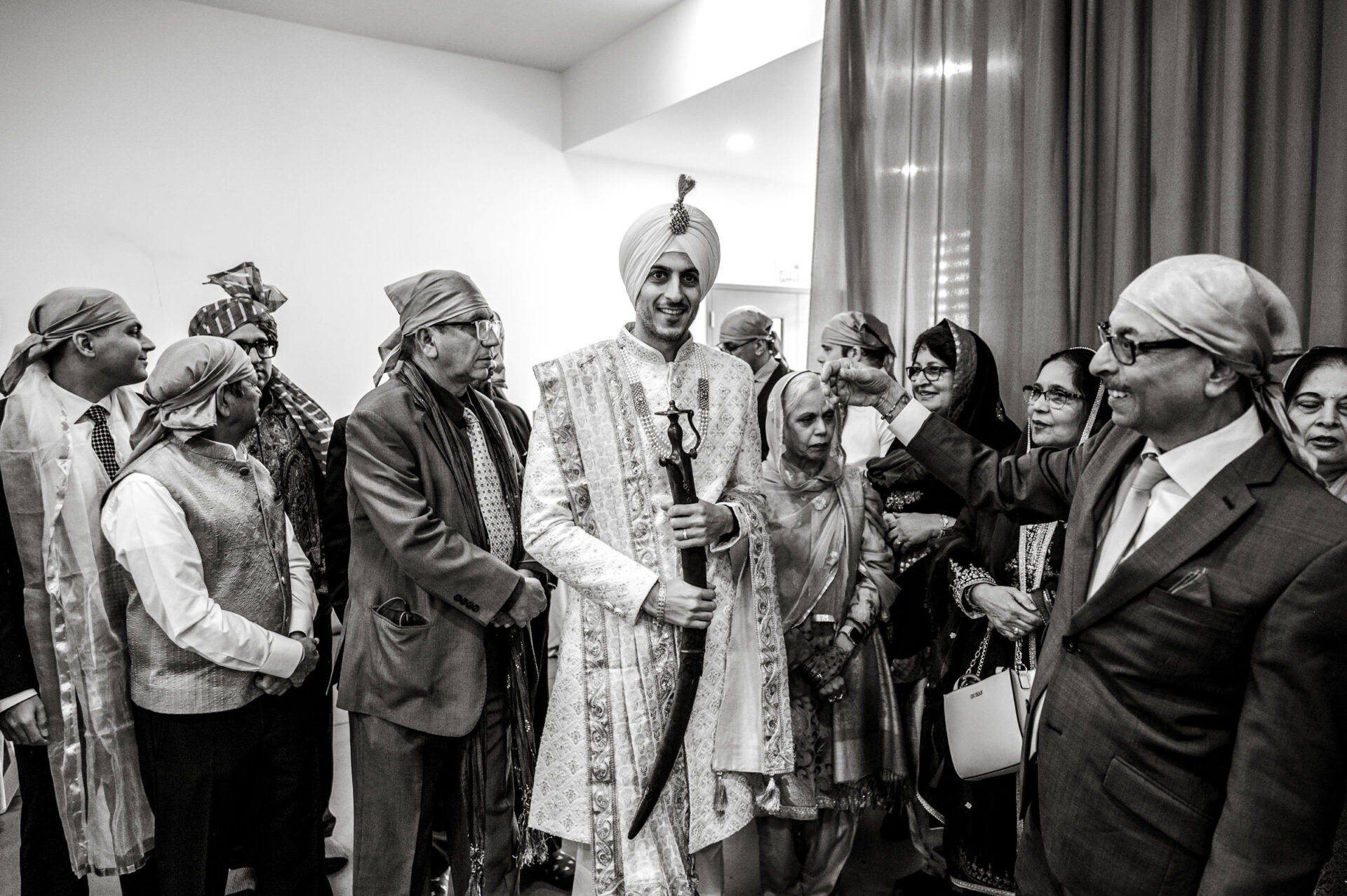 Groom in traditional attire holds a sword at a Sikh celebration, surrounded by smiling guests.