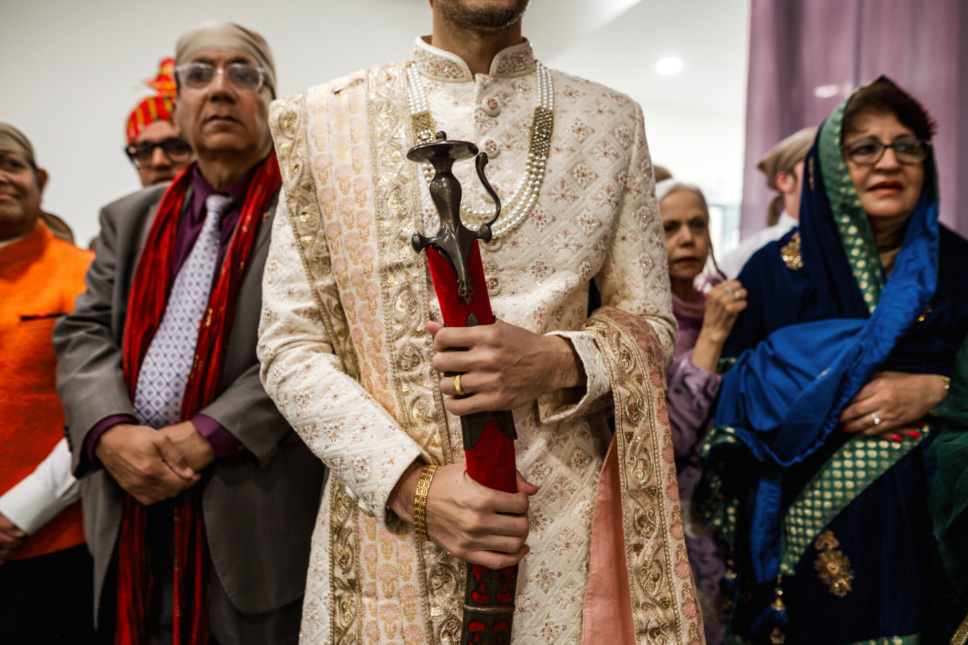 A Sikh groom in traditional attire holds a sword, surrounded by guests in colorful clothing.