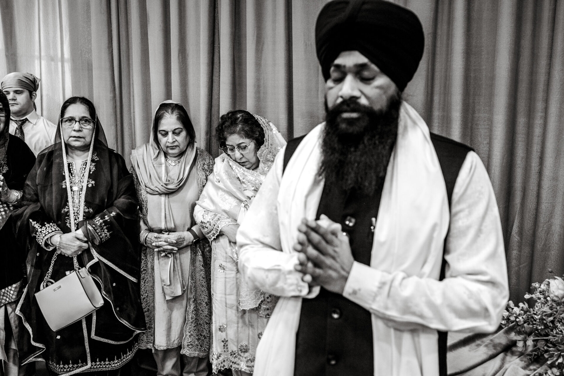 People at a prayer ceremony, one man clasping hands amid a Sikh wedding procession.