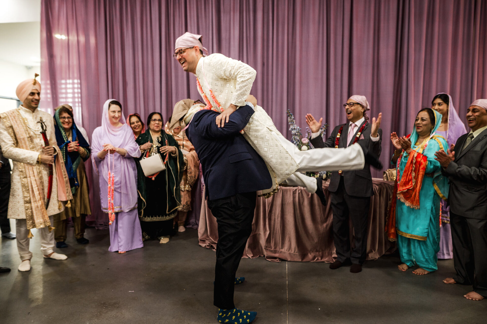 Amid a Sikh wedding, a person lifts another in joyful traditional attire.