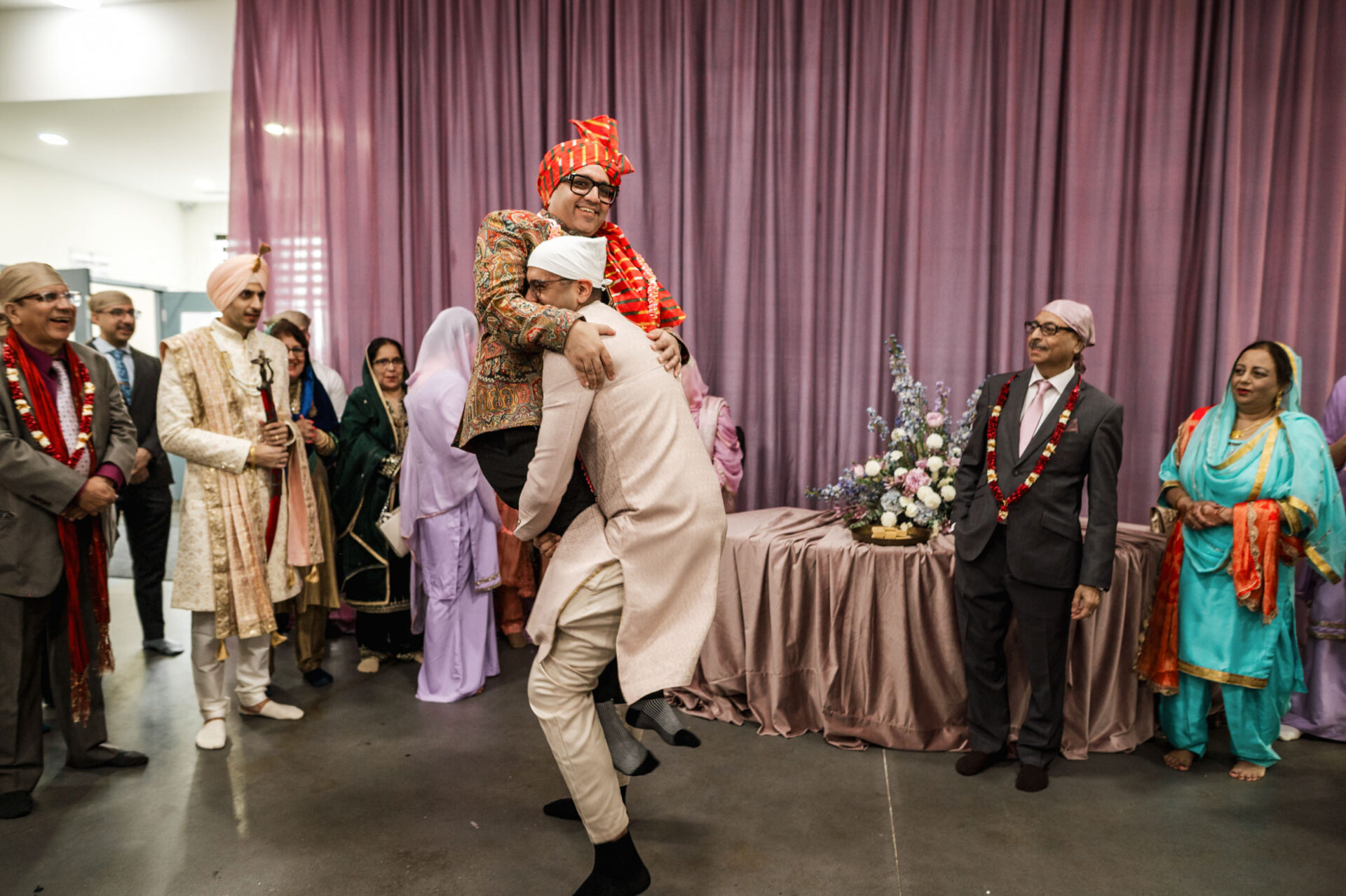 Two men laughing; one lifts the other at a joyful Sikh wedding with people watching.