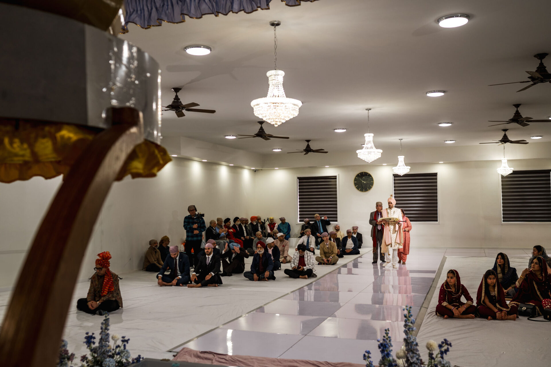 People gathered in a hall for a Sikh wedding, listening to a speaker at a podium.