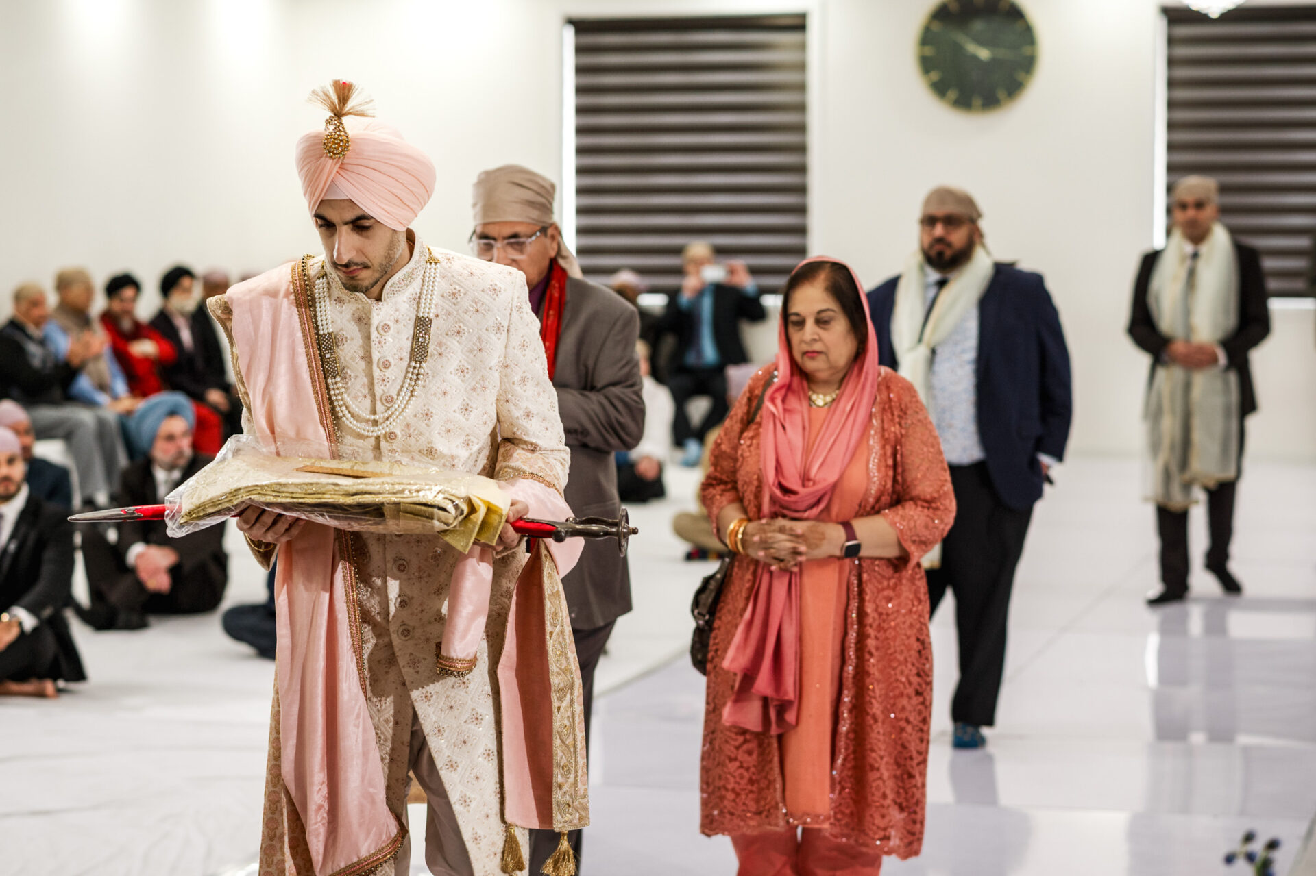 Person in traditional attire holds book during Sikh ceremony, surrounded by attendees in a hall.