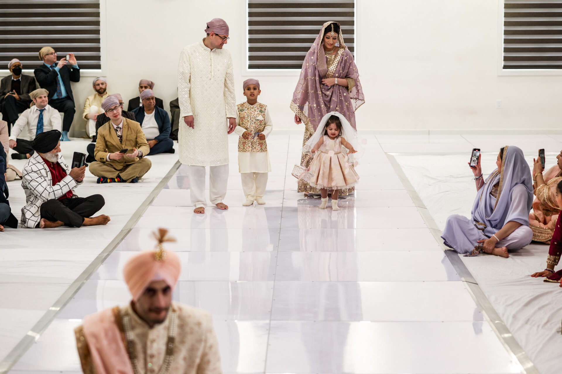 A family walks the aisle at a Sikh ceremony, surrounded by seated guests.